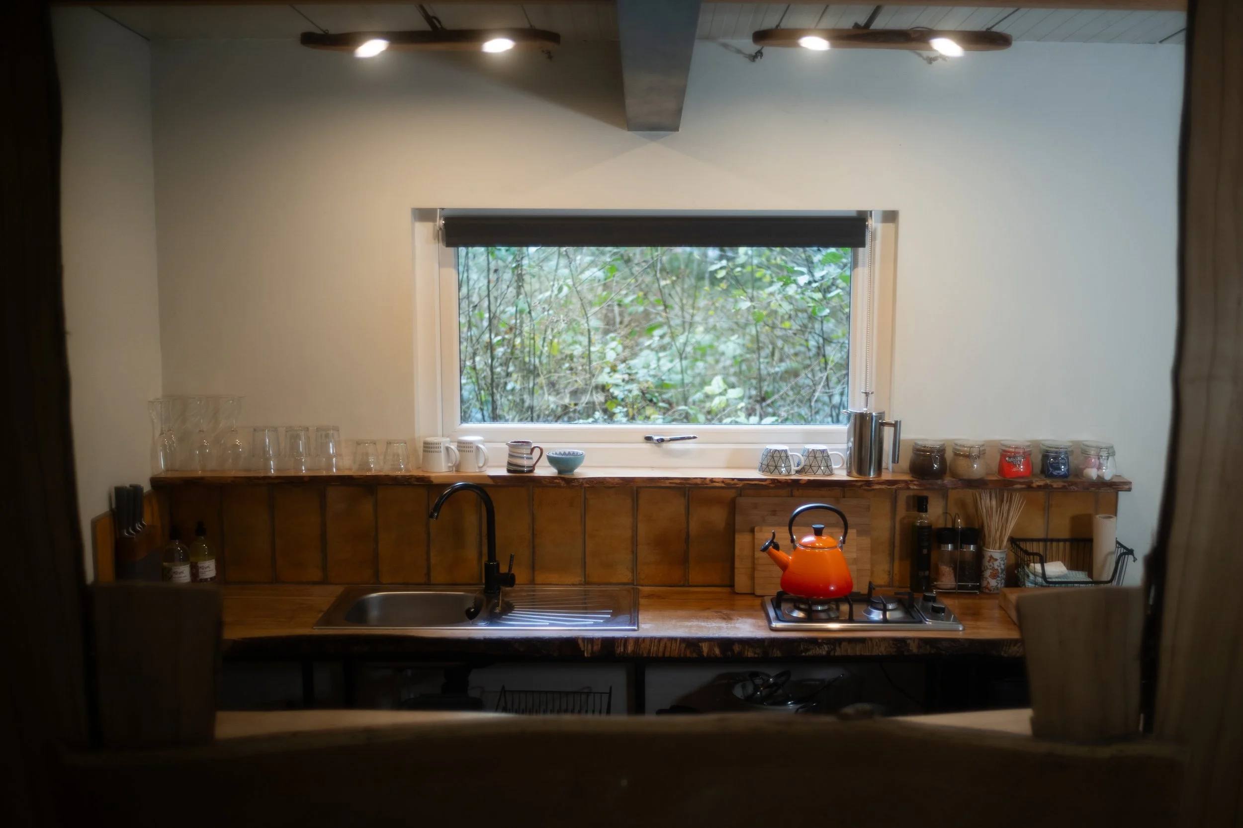 Wooden kitchen counter with a black faucet, orange kettle on a stove, various jars and utensils, a window with greenery outside, and overhead lighting.