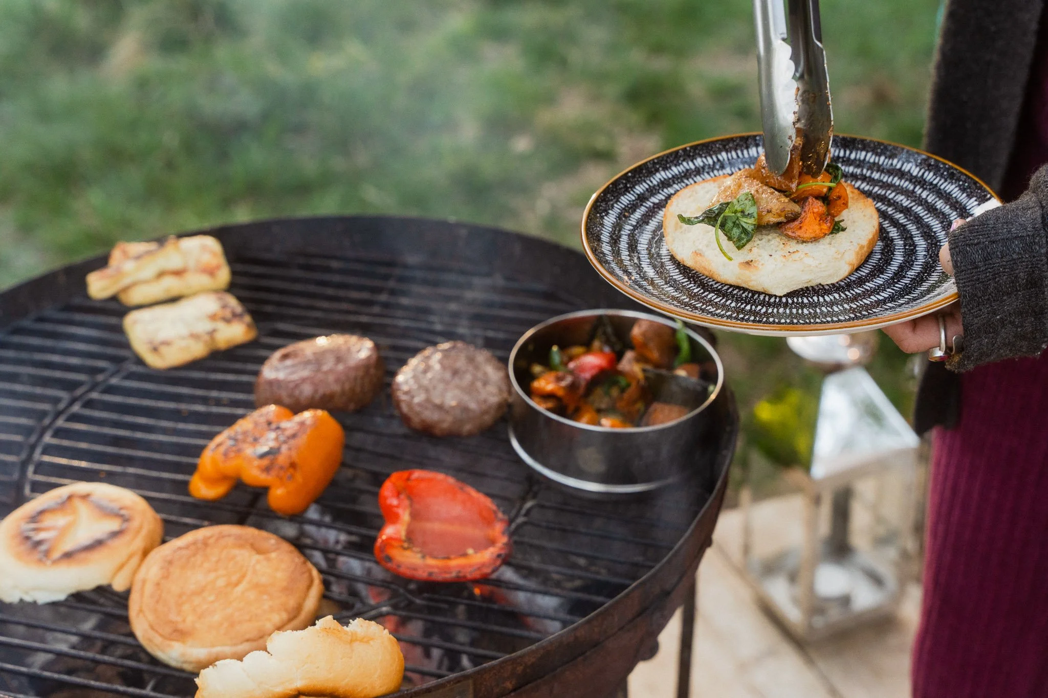 Person grilling assorted vegetables and hamburger patties on a barbecue grill, with someone placing a cooked piece of bread and vegetables on a plate.
