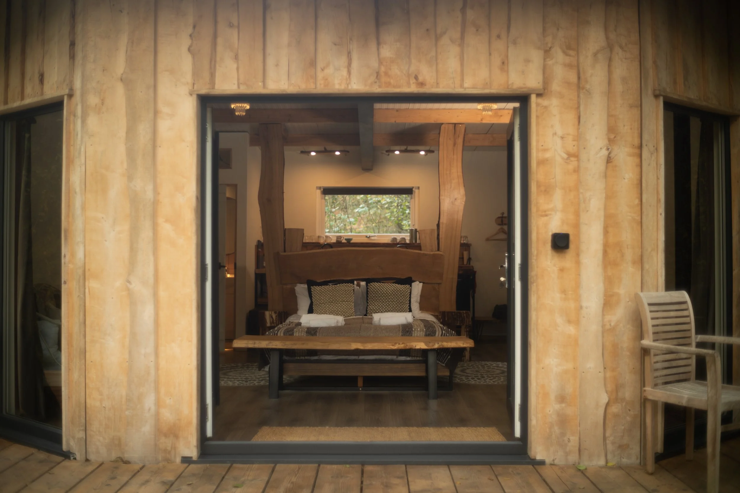 Interior view of the cosy bedroom at Brook with a large rustic wooden headboard, patterned cushions, and a small window overlooking a green outdoor scene, viewed through a sliding glass door from a wooden deck.