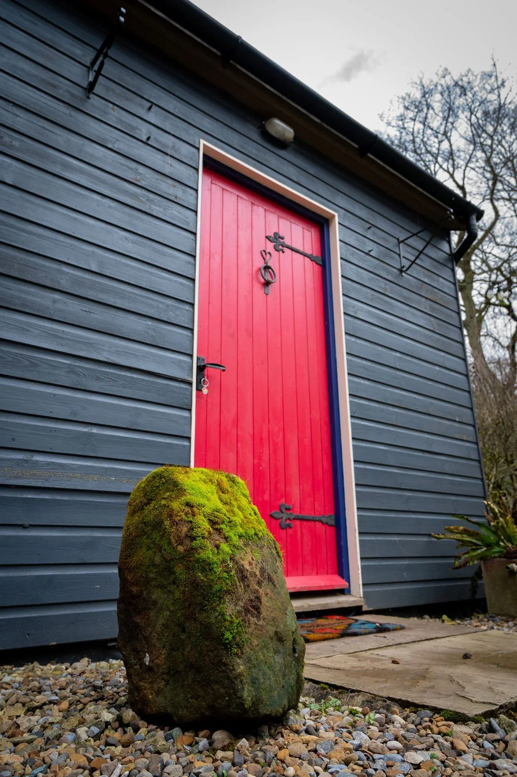 A red door with black decorative hinges on a dark blue wooden house, with a moss-covered rock in the foreground and trees in the background.