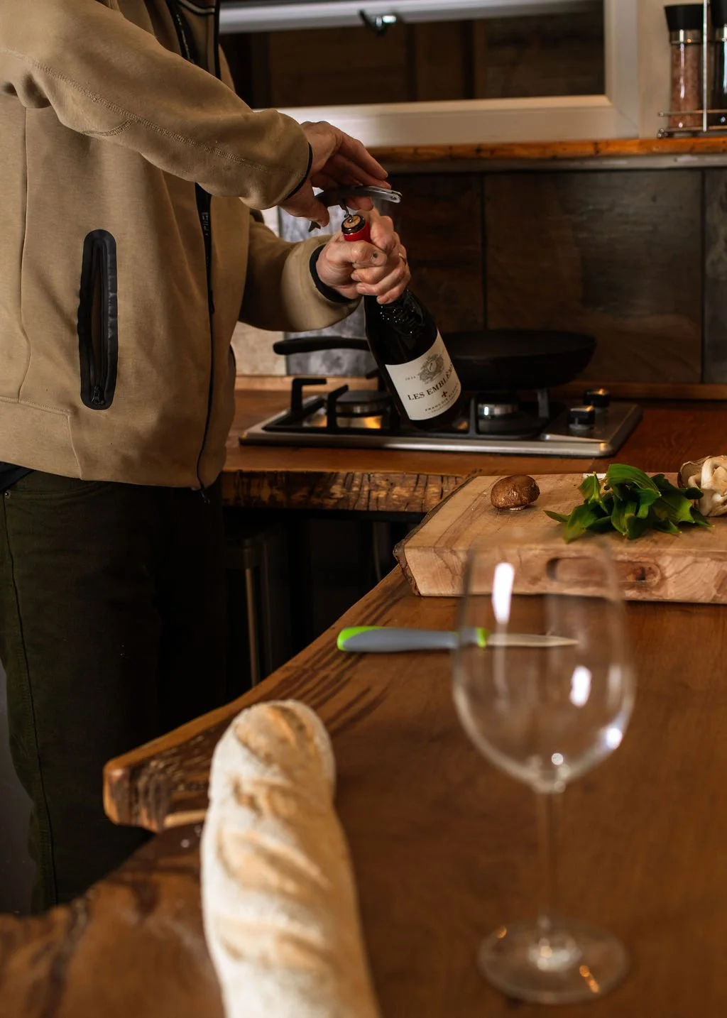 Person opening a bottle of red wine in a cozy kitchen with a wooden countertop, fresh mushrooms, herbs, a bread loaf, and an empty wine glass.