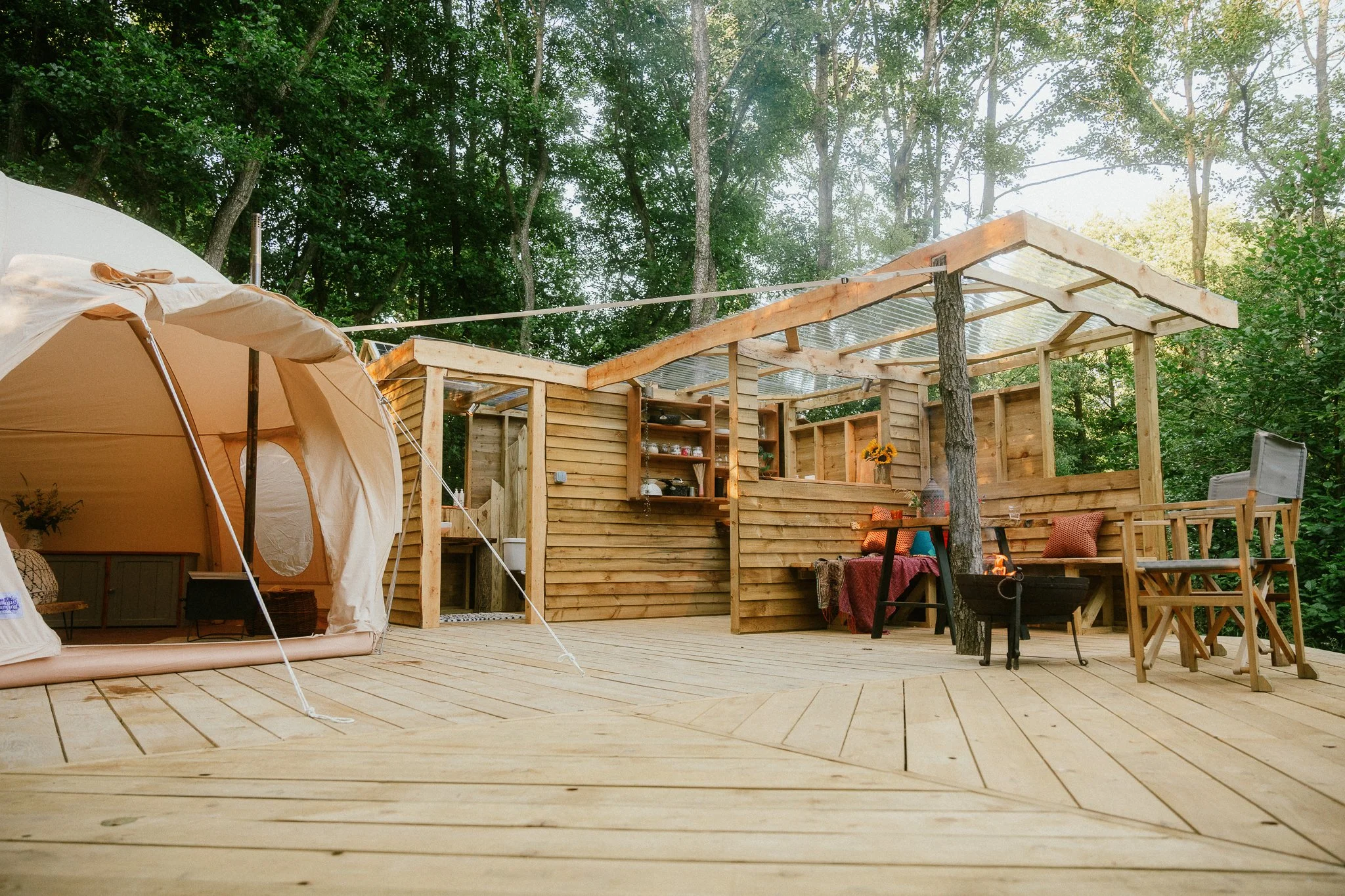 A wooden outdoor deck with a beige tent on the left, and a wooden structure with seating, a fire pit, and open shelving in the background, surrounded by trees.