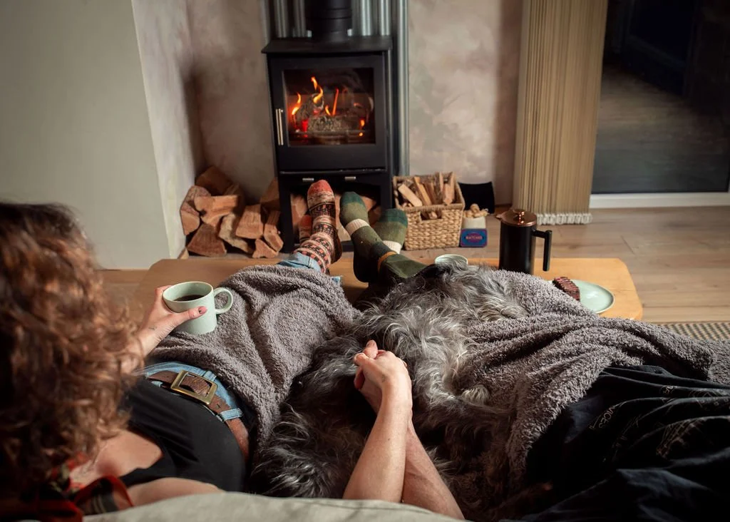 Two people relaxing on a couch by a fireplace, one holding a mug, with a dog lying on their lap.