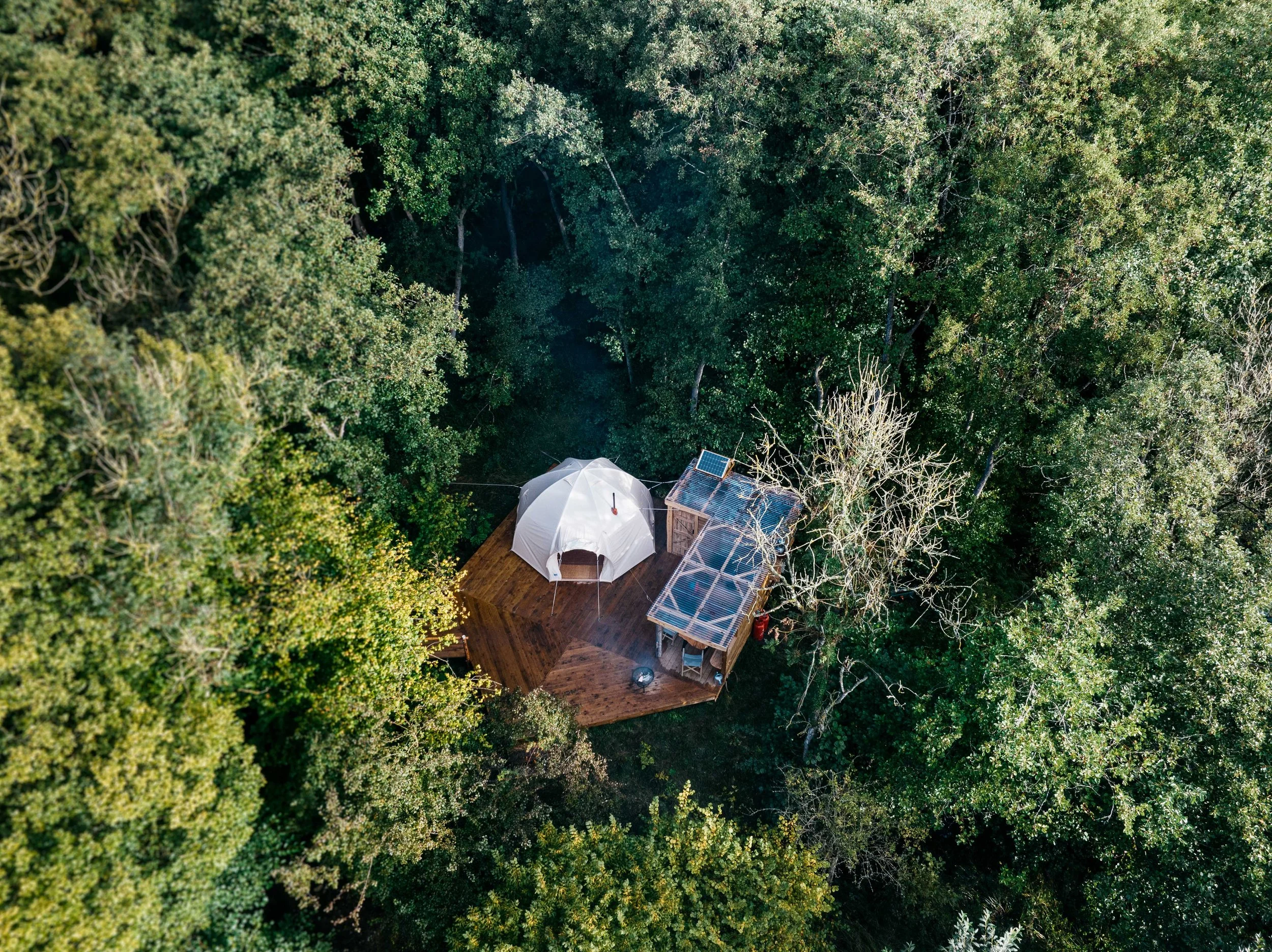 Aerial view of a tiny house with solar panels and an umbrella on a wooden deck, surrounded by dense green trees in a forest.