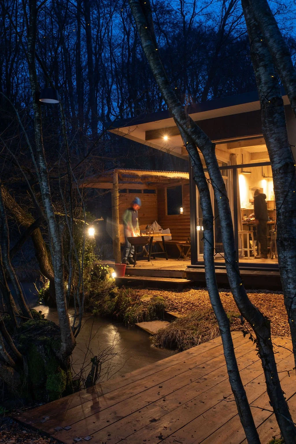 A cozy lit cabin in a wooded area at dusk with people inside and outside, including one person grilling by the porch and another inside preparing food.