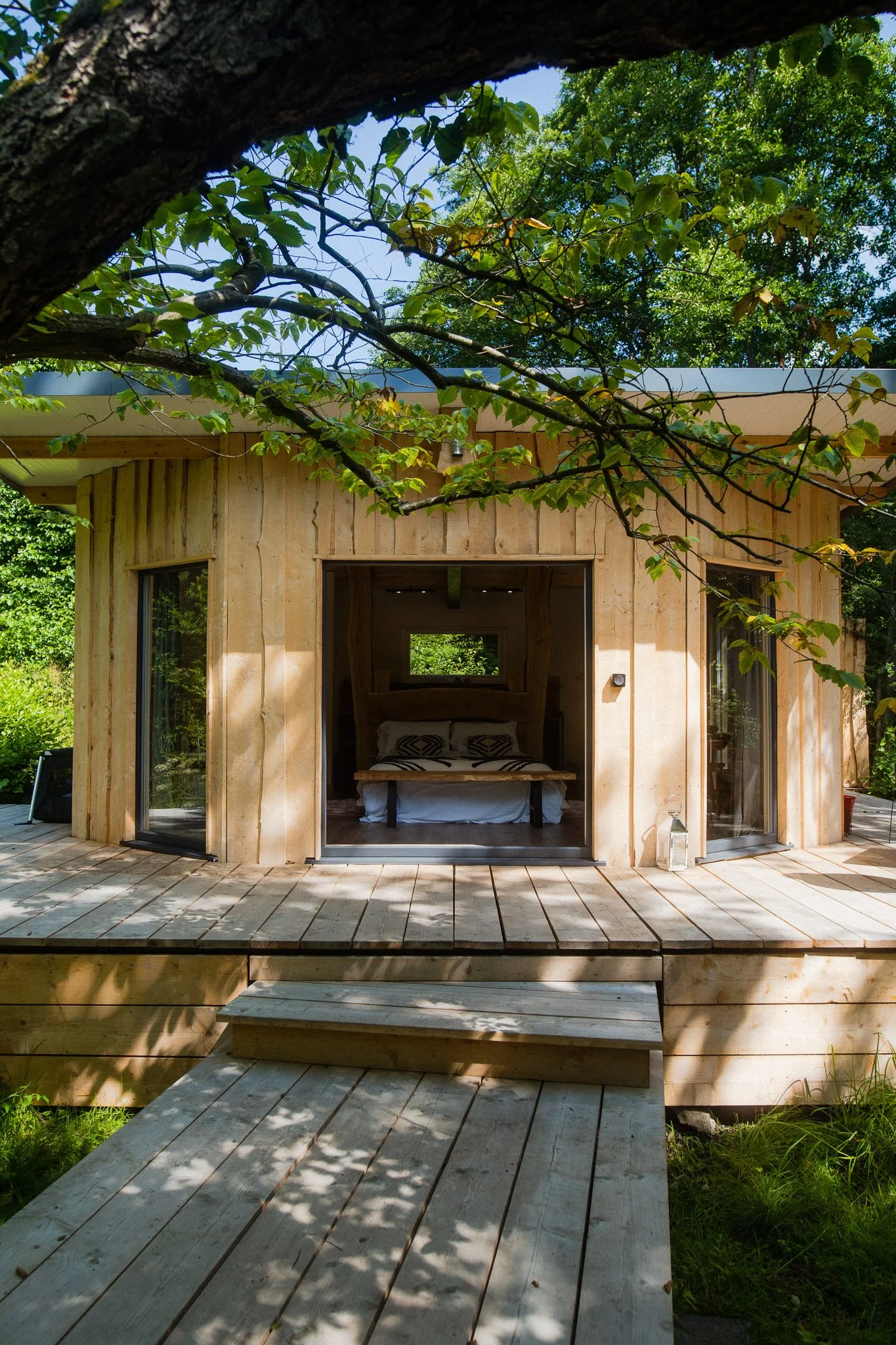 A small, round wooden cabin with large glass sliding doors, set in a lush green forest. The cabin has a porch with wooden steps leading up to it, and inside, a bed with patterned pillows is visible.