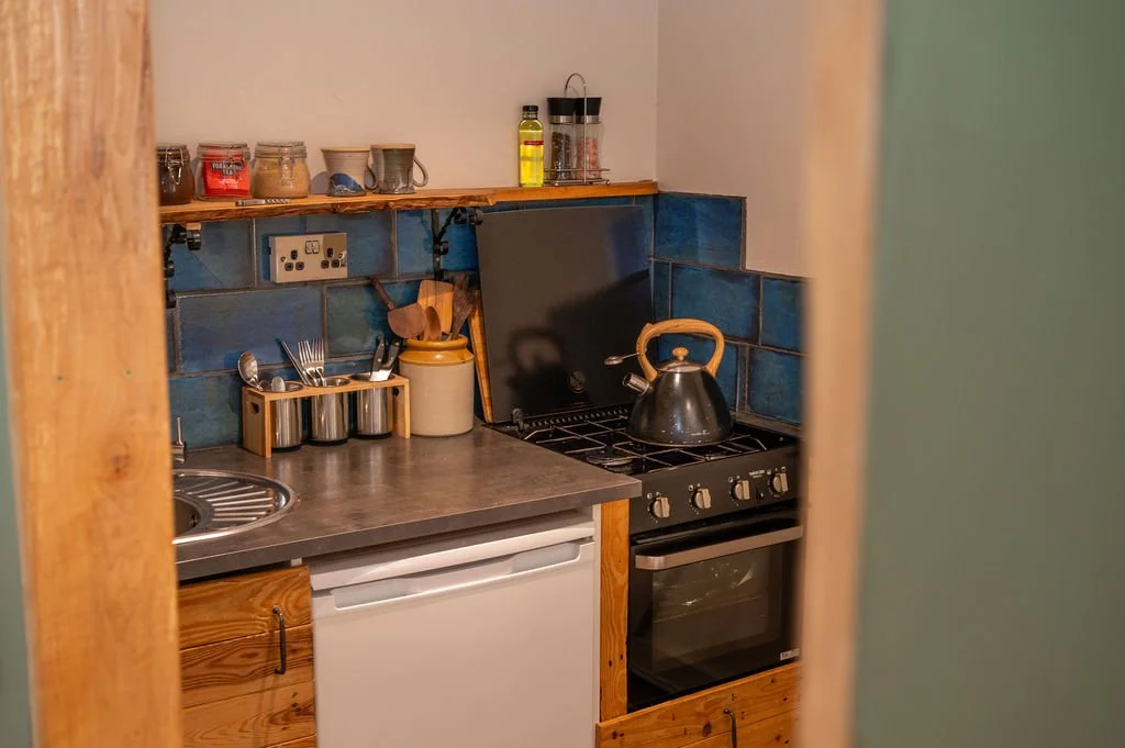 A small kitchen with blue tiled backsplash and wooden cabinets; contains a stove with a kettle, a small fridge, and a countertop with utensils, jars, and bottles.