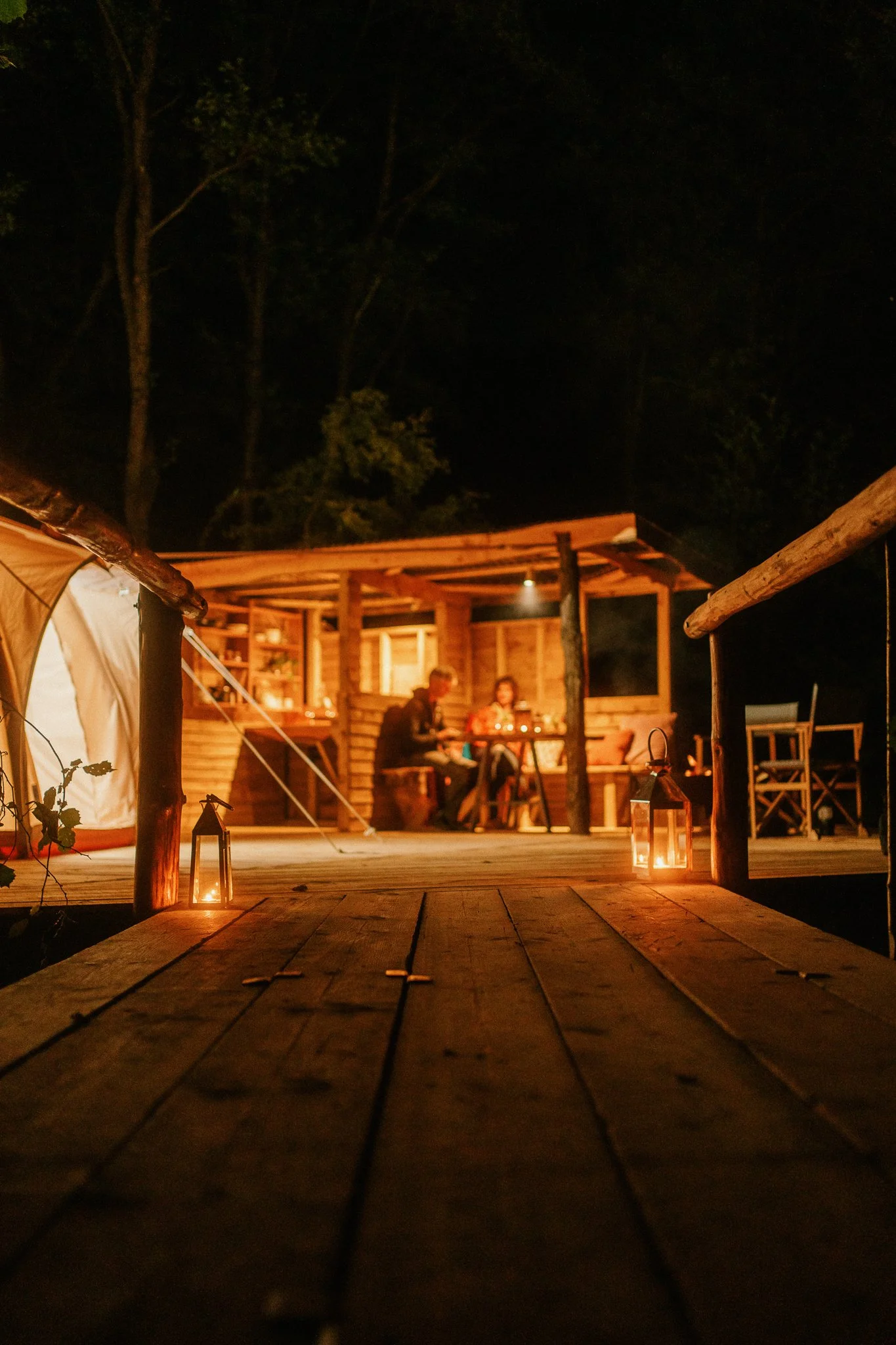 Nighttime scene of people dining in a wooden cabin with lanterns on the deck and a tent to the side, surrounded by trees.