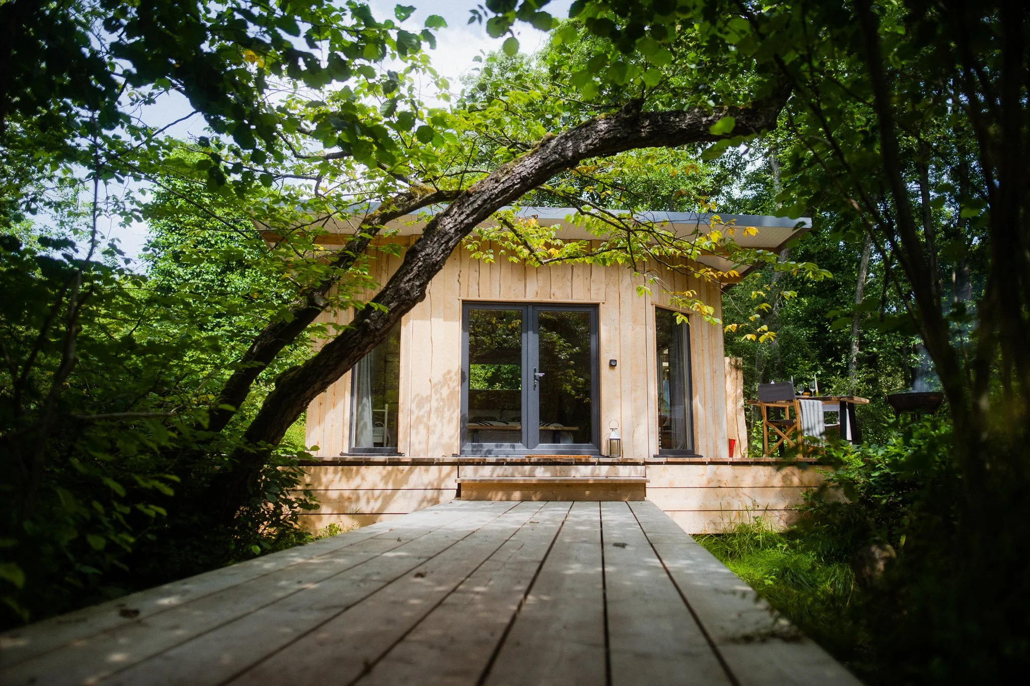 A small wooden cabin with large glass doors and windows, surrounded by lush green trees, with a wooden walkway leading to the entrance.