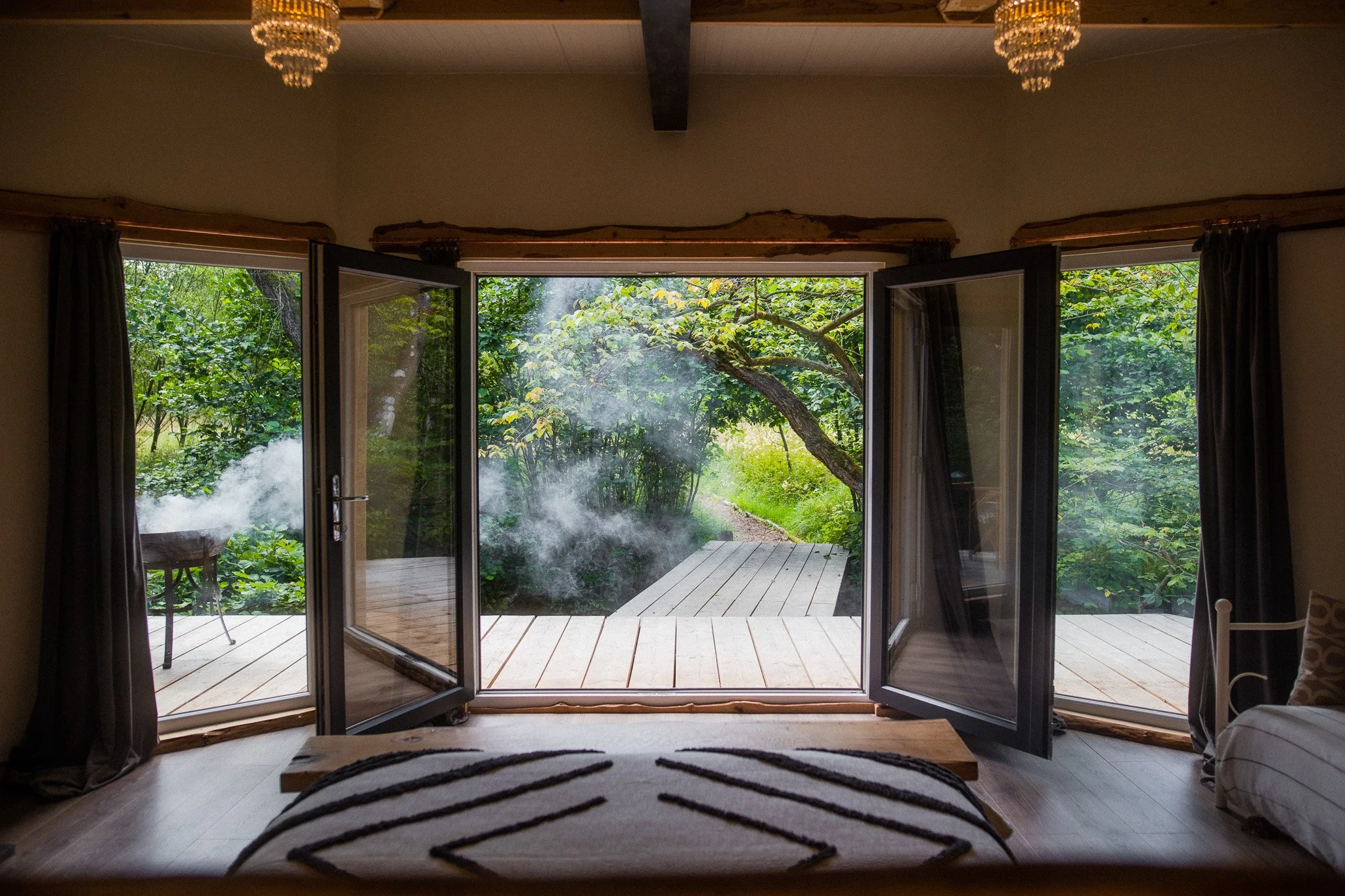 Inside a cozy room with open glass doors leading to a wooden deck and a lush green garden with trees and bushes outside.