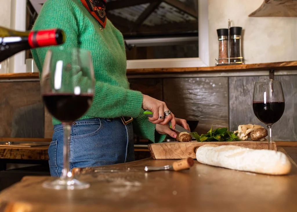 A person wearing a green sweater and jeans slicing mushrooms on a wooden cutting board in a rustic kitchen with two glasses of red wine on the counter.