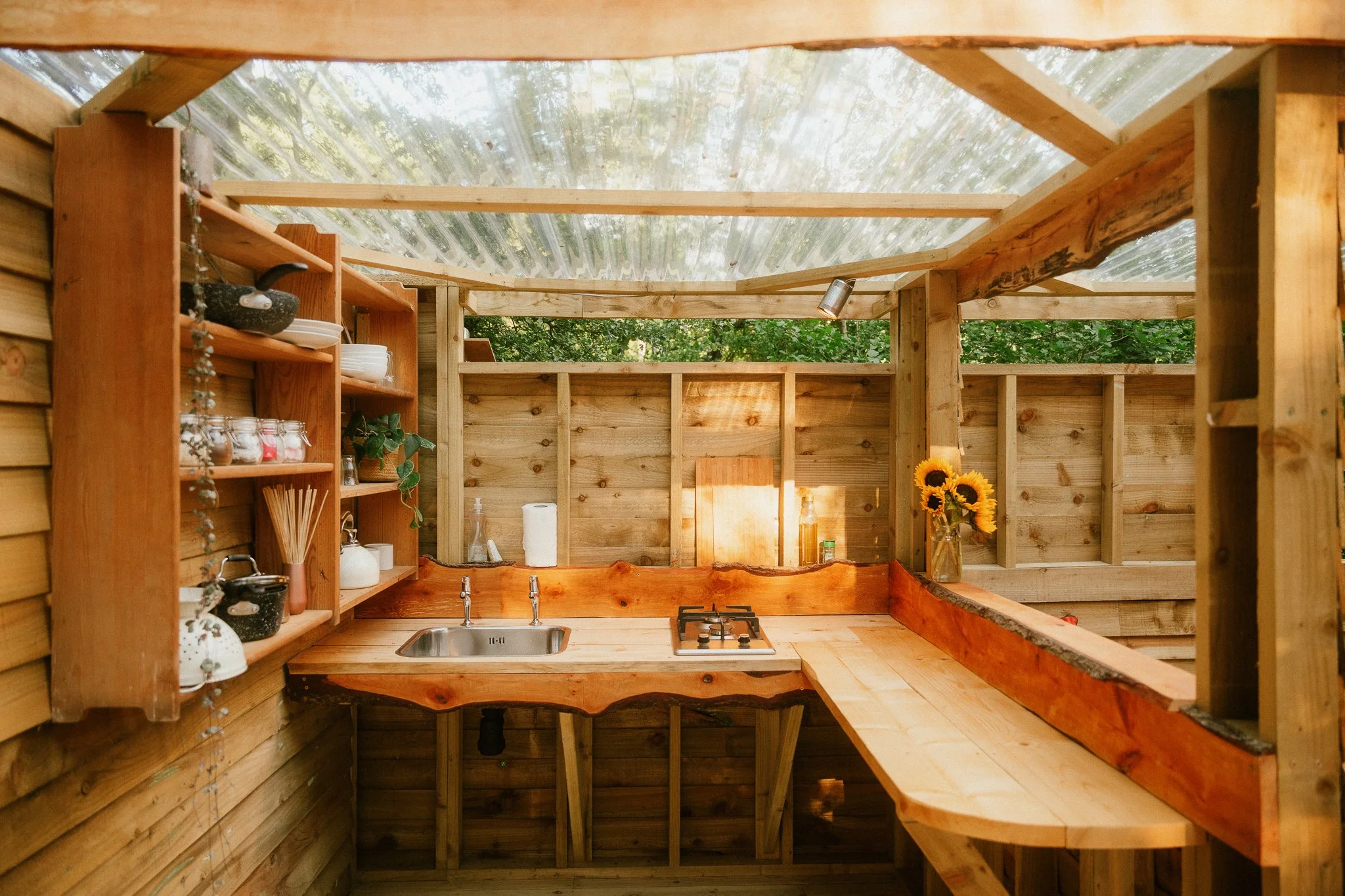 Interior of a rustic outdoor kitchen with wooden countertops, shelves, a small sink, a stove, and a vase of sunflowers, under a transparent roof with surrounding wooden walls.