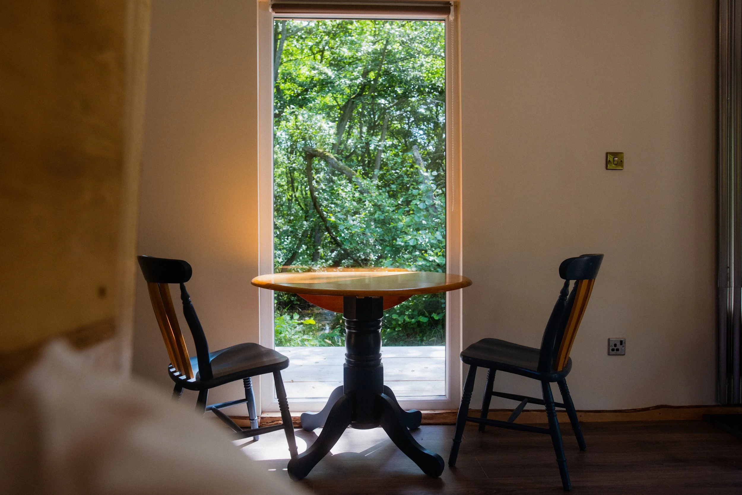 A small round wooden table with two dark wooden chairs located in front of a large window showing green trees outside.