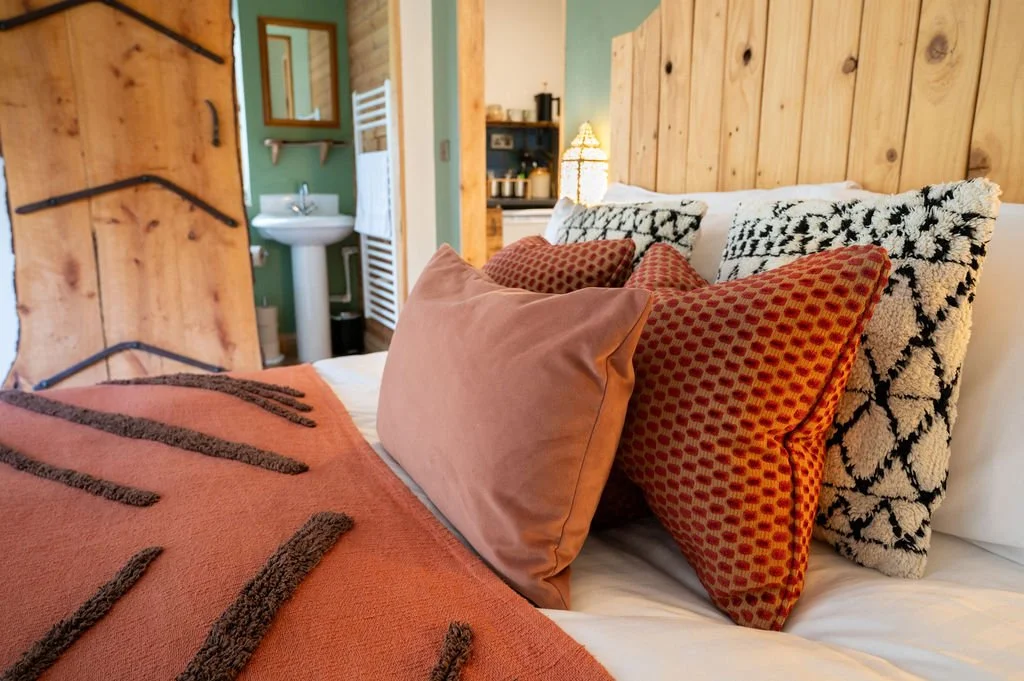 A cozy bedroom with a bed featuring multiple decorative pillows, a wooden headboard, and a rustic theme. In the background, there's a bathroom with a sink and a mirror.