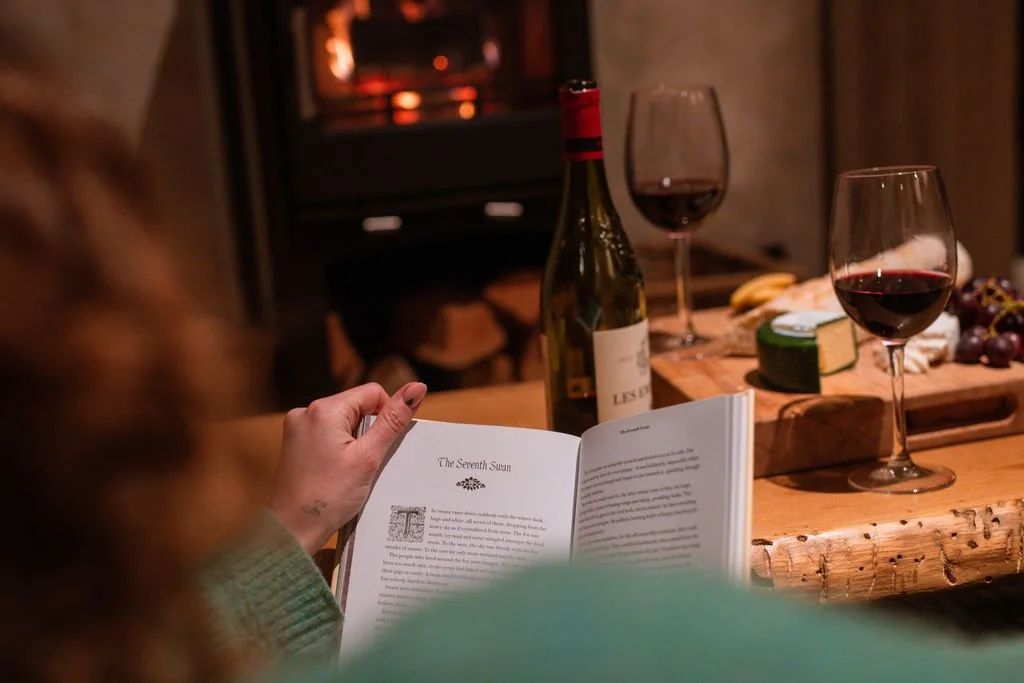 Person reading a book titled "The Seventh Sun" at a cozy dinner table with two glasses of red wine, a bottle of wine, cheese, and grapes, in front of a fireplace.