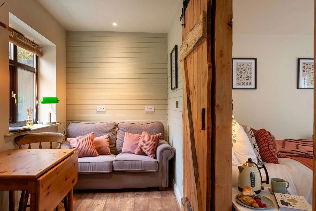 A cozy living area with a gray sofa adorned with pink and patterned throw pillows, next to a window with a bamboo blind, a wooden table, and a chair. A sliding barn door separates the living space from a bedroom, where a bed with a teapot, cup, croissants, and a magazine are visible.