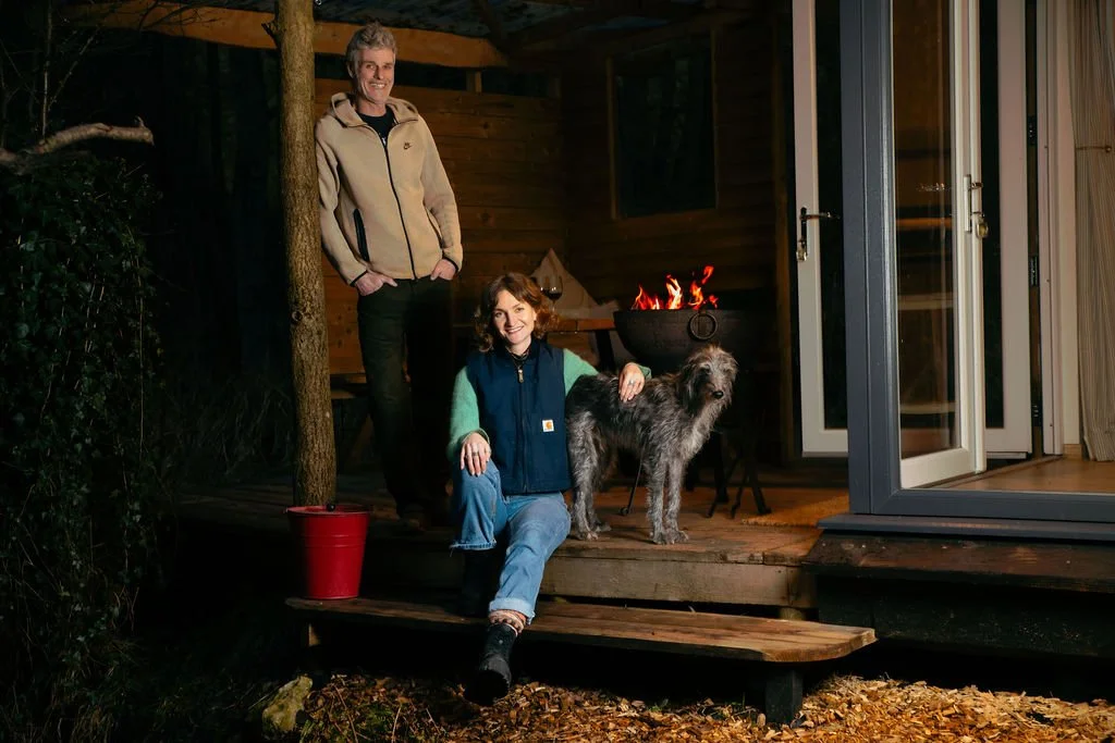 Owners, Katy and Skot, sitting on the deck of the wooden cabin at night with a fire and dog