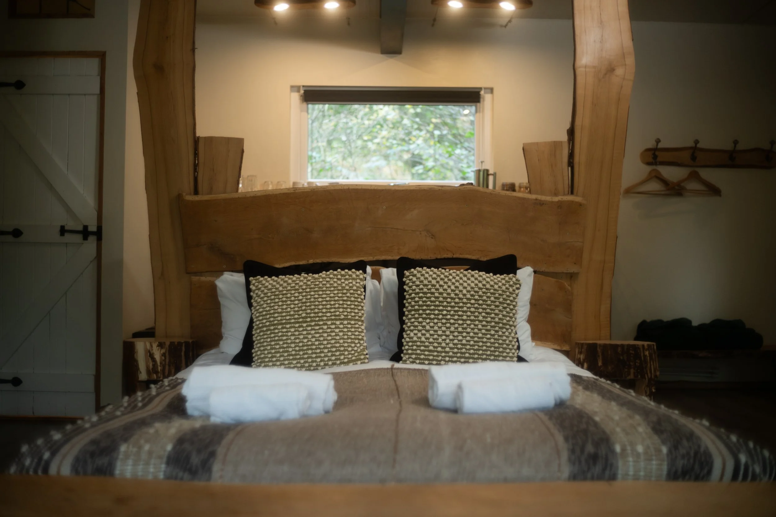 A cozy bedroom with a wooden bed and headboard, two decorative pillows, and neatly folded white towels on the bed. A window with a view of trees is behind the bed.