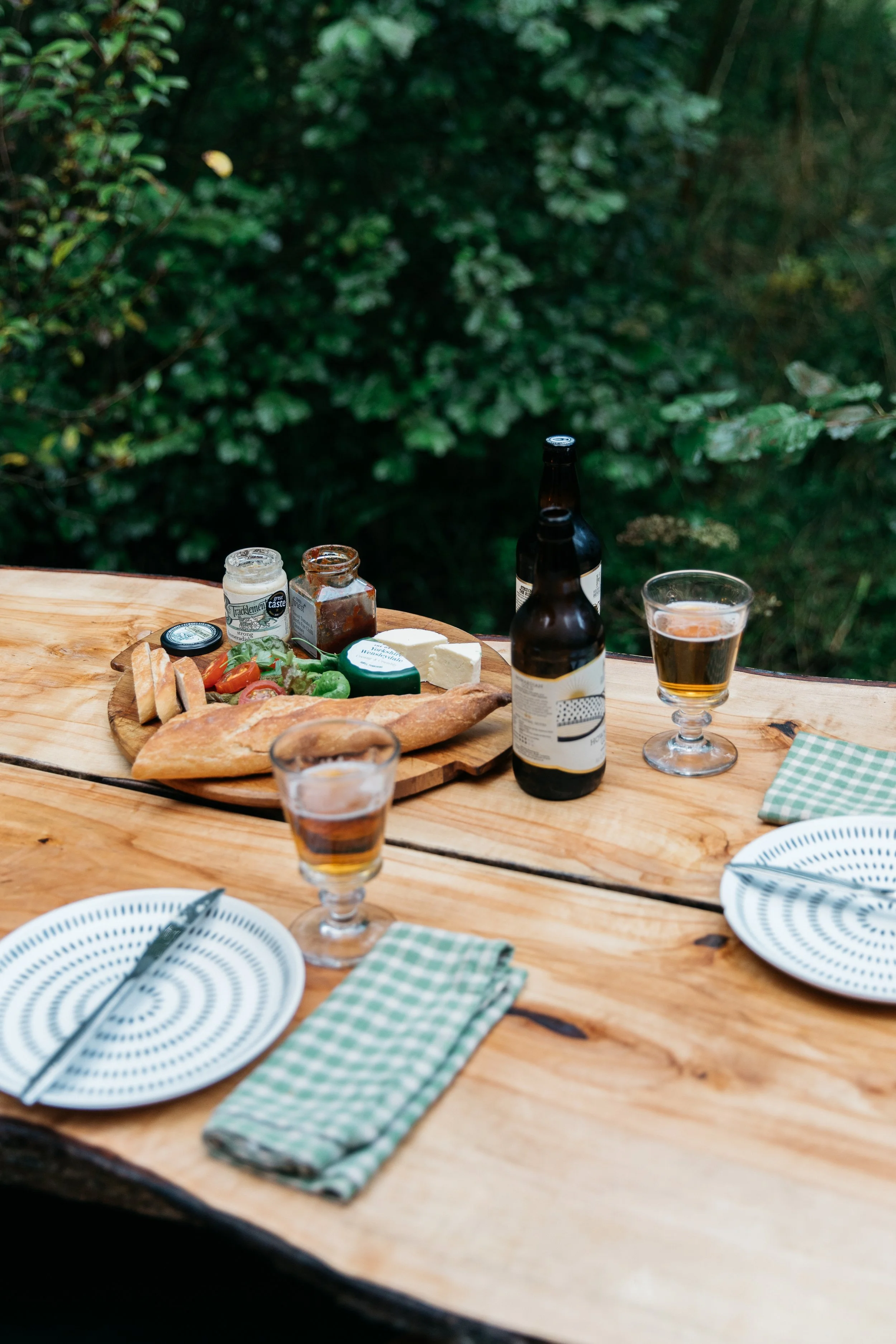 Outdoor dining setup with a rustic wooden table, two plates with cutlery and green checkered napkins, two glasses of beer, a cheese platter with bread, cheese, vegetables, and condiments, and a forest background.