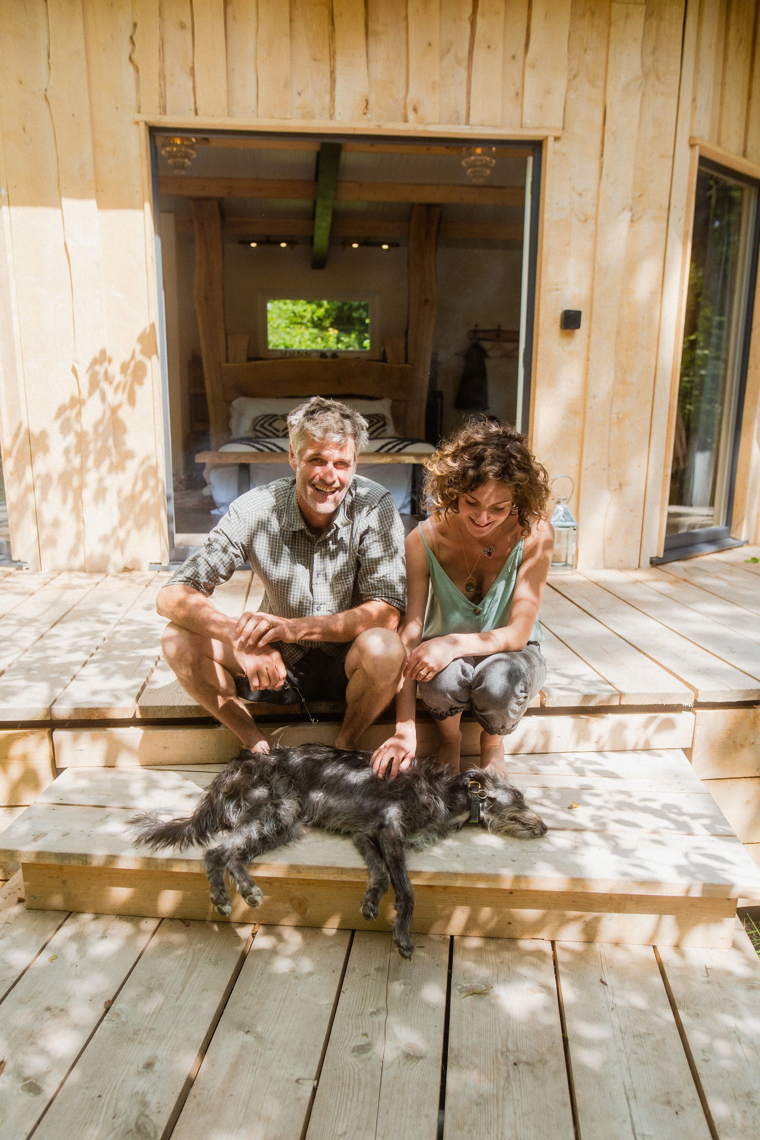 Owners, Katy and Skot, sitting on the stoop of their hand-built wooden cabin, smiling at the camera, with a dog lying in front of them.
