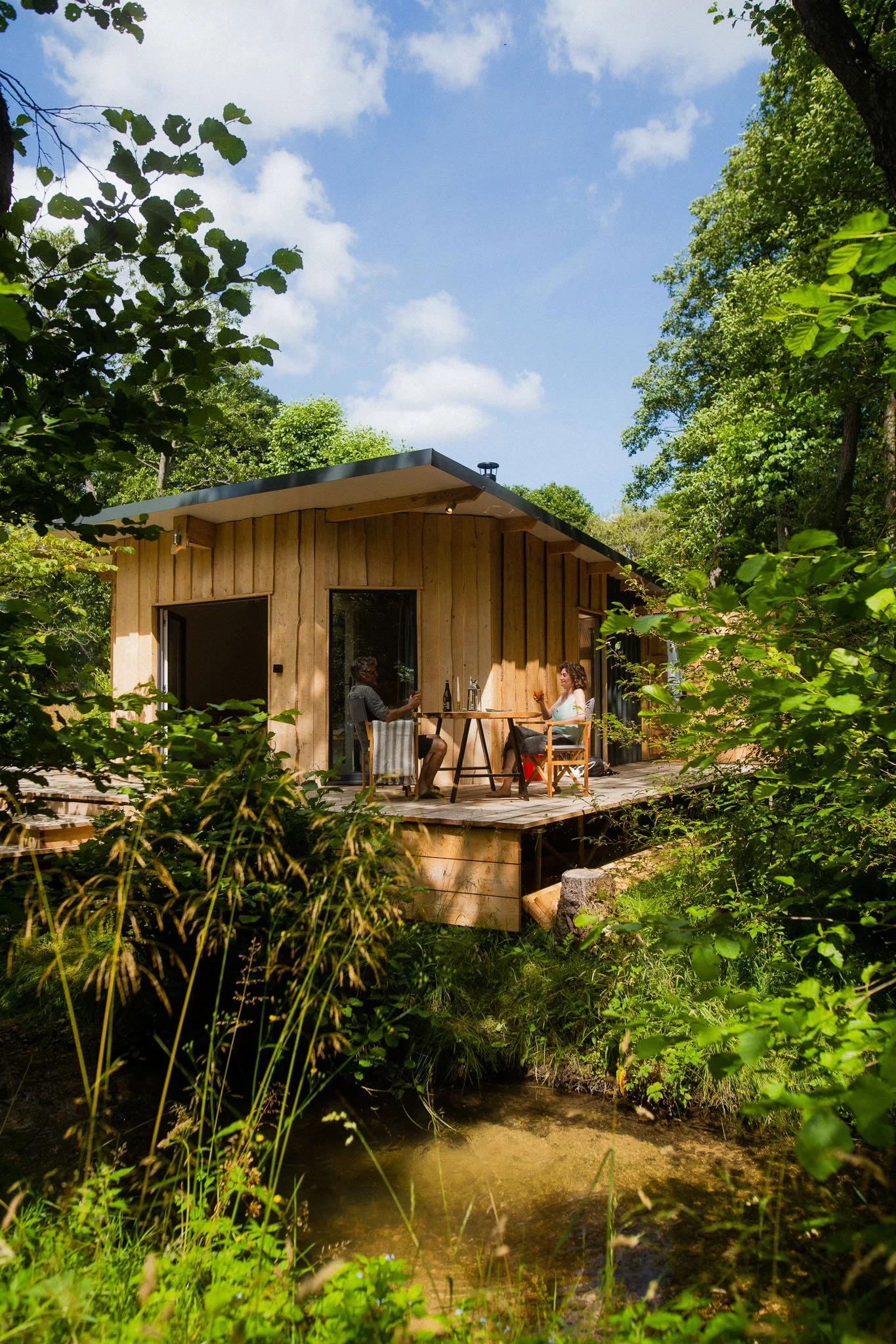 A wooden cabin on a deck surrounded by green trees, with two people sitting at a table drinking and talking on a sunny day near a small stream.