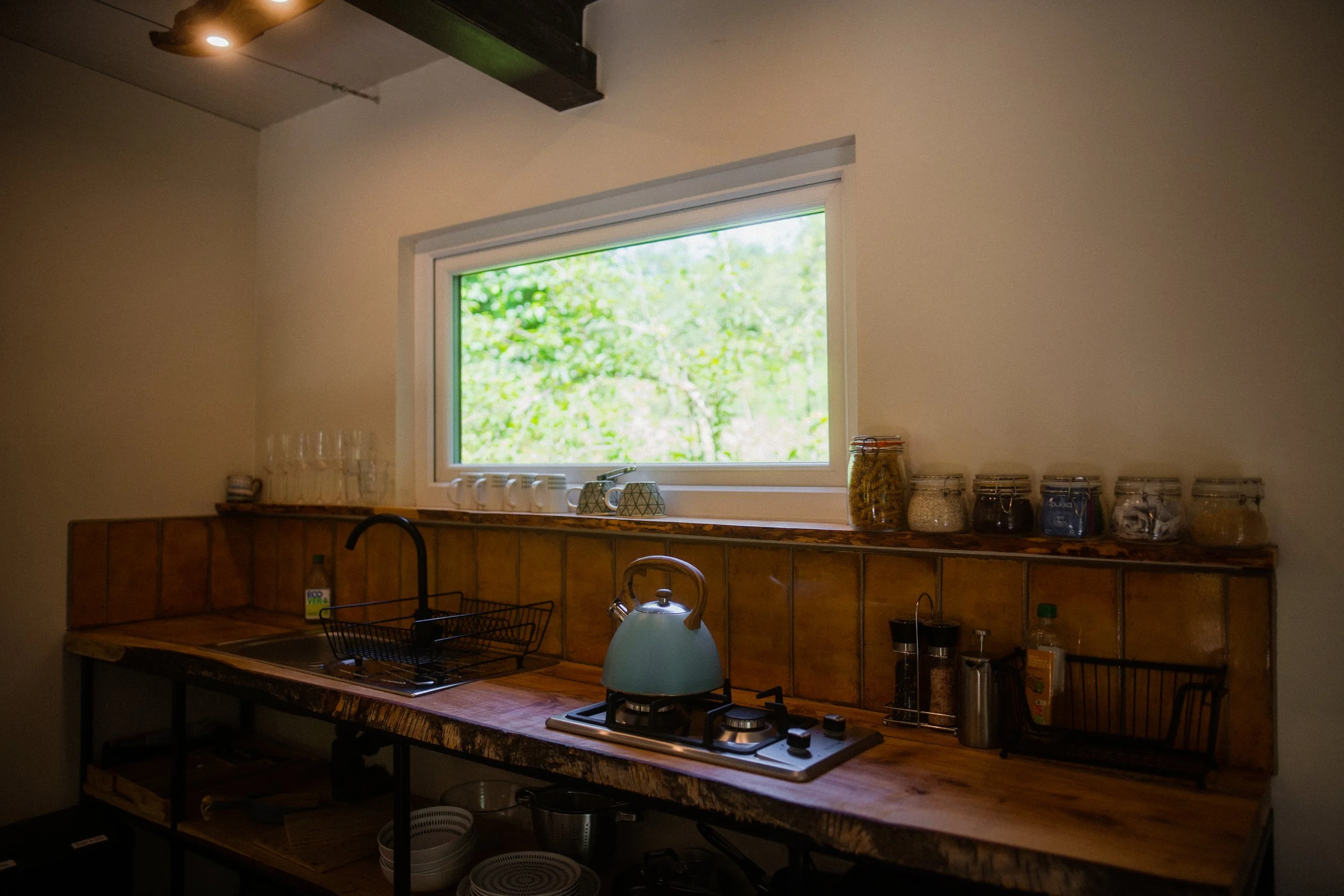 Kitchen with a wooden countertop, black faucet, tea kettle, and jars of ingredients, with a large window showing greenery outside.