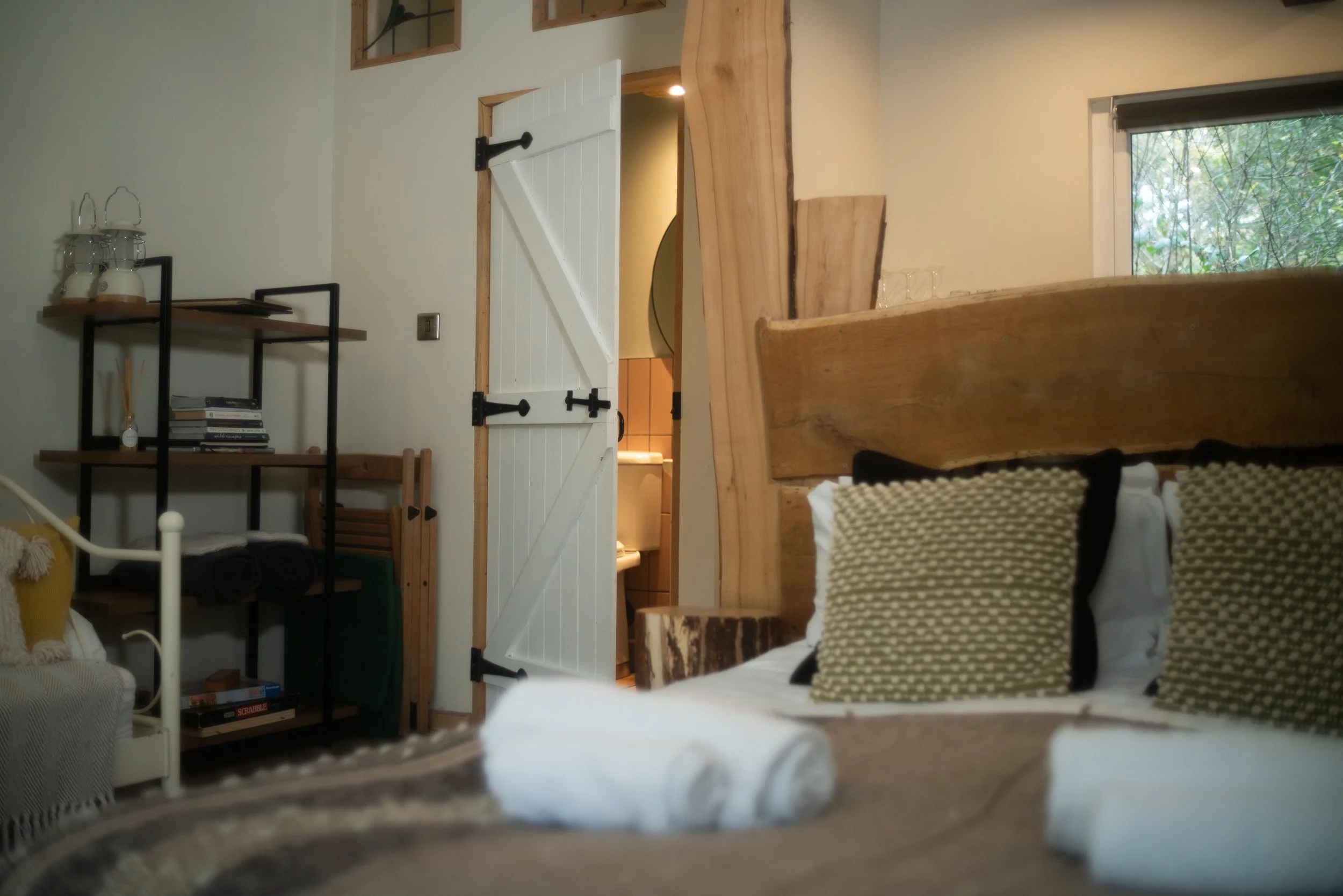 A cozy bedroom with a wooden headboard, patterned pillows, a window showing greenery outside, a white door leading to a bathroom, and a black metal shelf with books and decorative items.