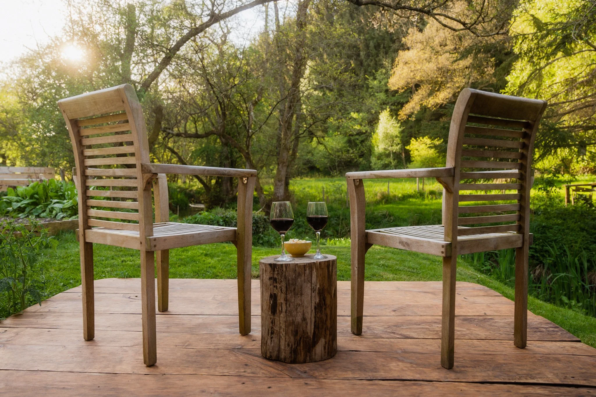 Two wooden chairs facing each other with a small tree stump table in between. On the table are two glasses of red wine and a bowl of cheese. The scene is set outdoors on a wooden patio with green grass and trees, and the sun is shining through the trees.