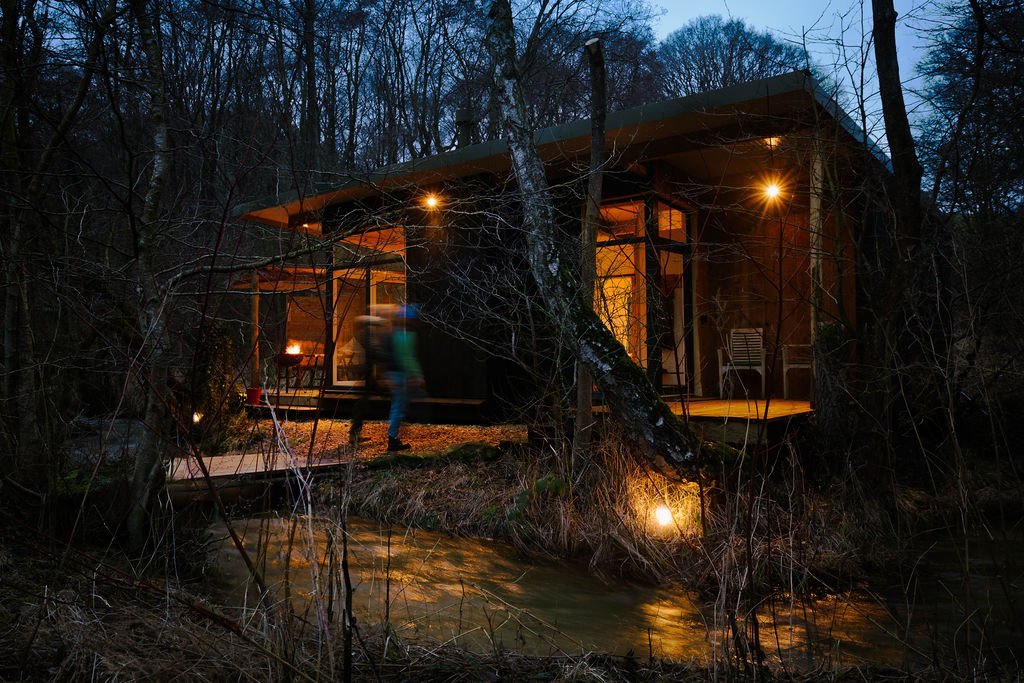 Beck by the stream in the wooded area at dusk, with warm interior lights shining through large windows . 