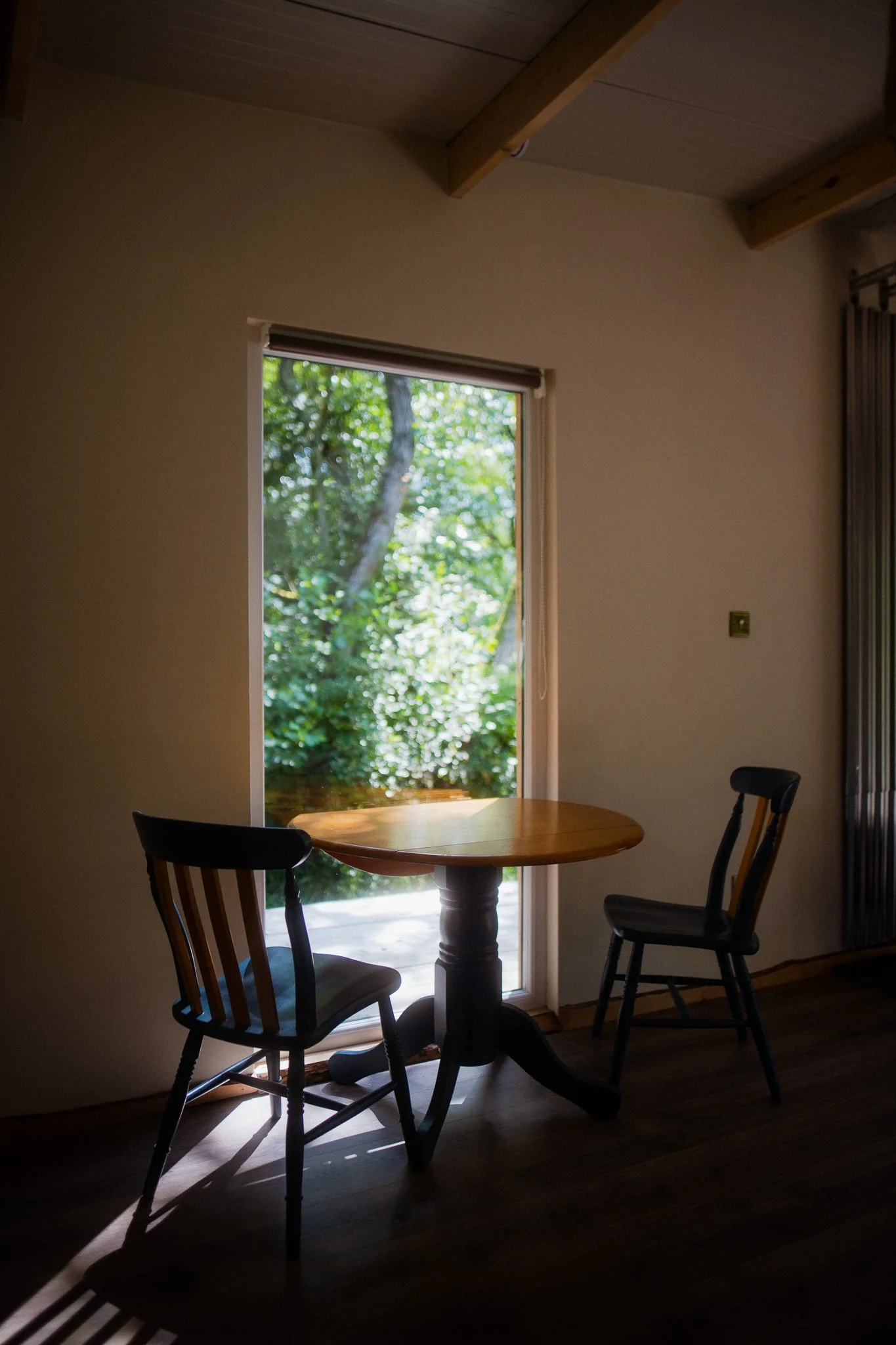 Interior of a room with a small round wooden table and two chairs, backlit by a large window showing greenery outside