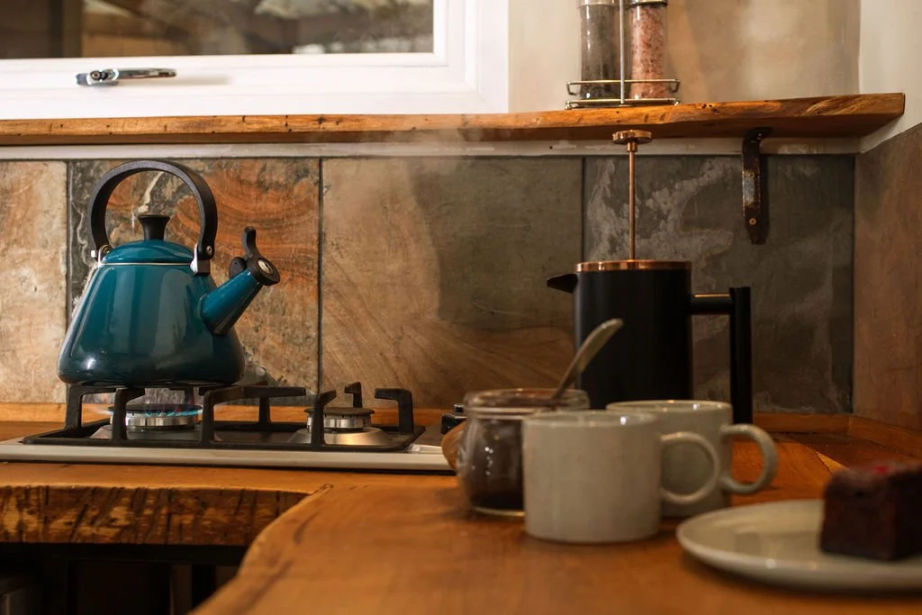 Kitchen countertop with a teal kettle on the stove, a jar of jam, two mugs, a plate with a piece of cake, and a French press coffee maker, with a window and wooden shelf in the background.