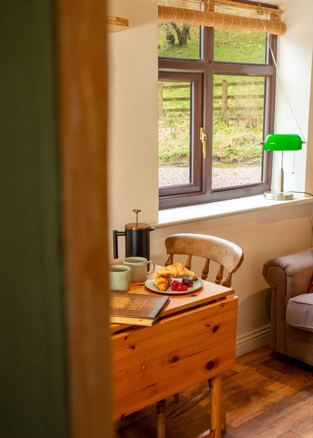 A cozy interior of a room with a wooden side table holding a breakfast plate with croissants and berries, two green mugs, a French press, and a book. A window with a bamboo shade reveals a grassy outdoor scene. There is a green desk lamp and part of a beige armchair in the room.