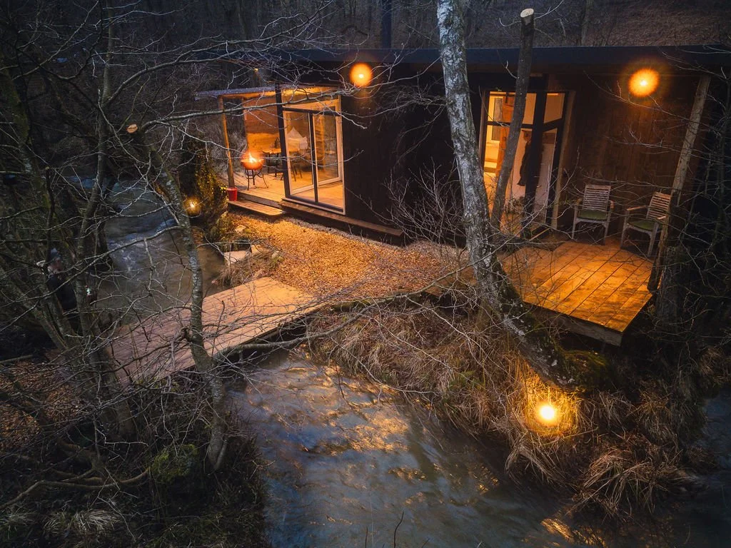 A treehouse built into trees over a river at night, illuminated by warm outdoor lighting, with a small wooden deck and large glass doors leading inside.