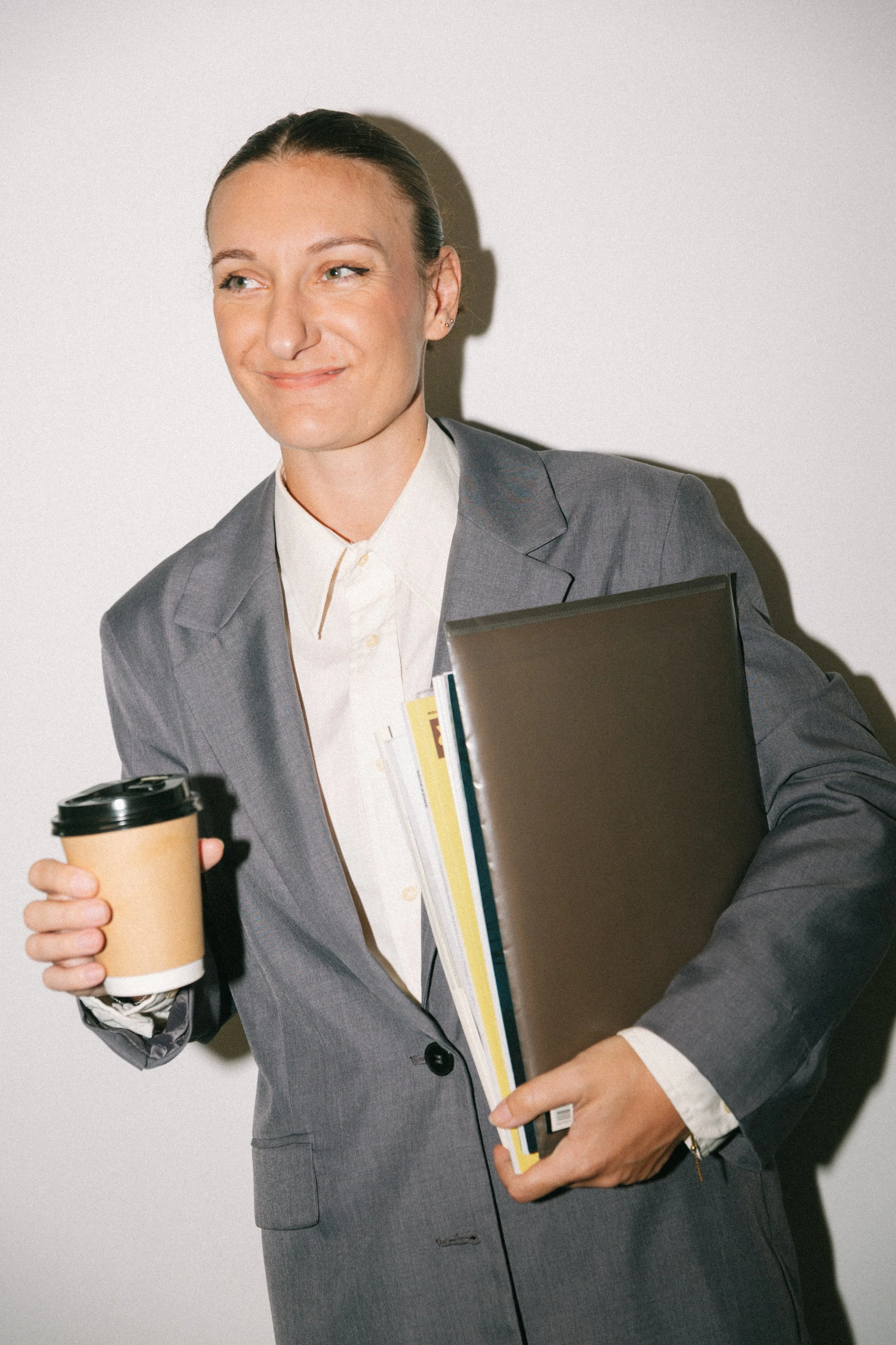 "Femme d'affaires souriante portant un costume gris, tenant un café à emporter dans une main et un classeur avec dossiers dans l'autre, en présence d'une feuille volante." je garde cette description auto générée, j'aime bien