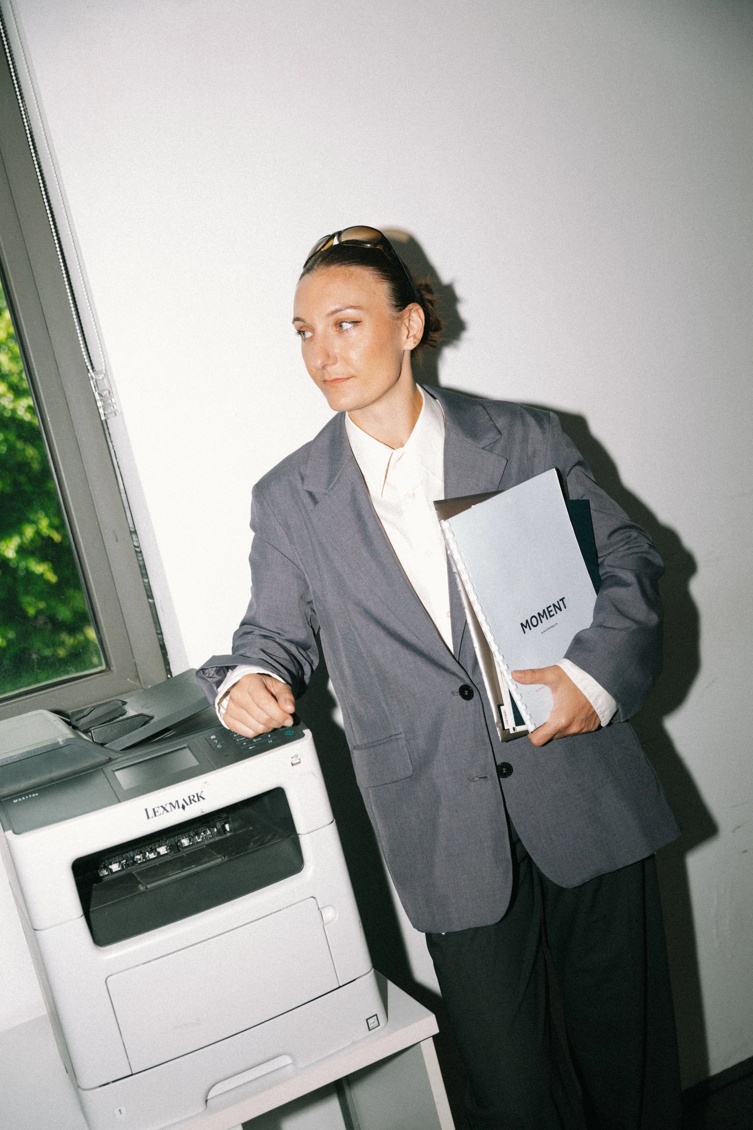 Une femme d'affaires en costume gris utilisant une photocopieuse dans un bureau lumineux, tenant un dossier intitulé 'Moment'.