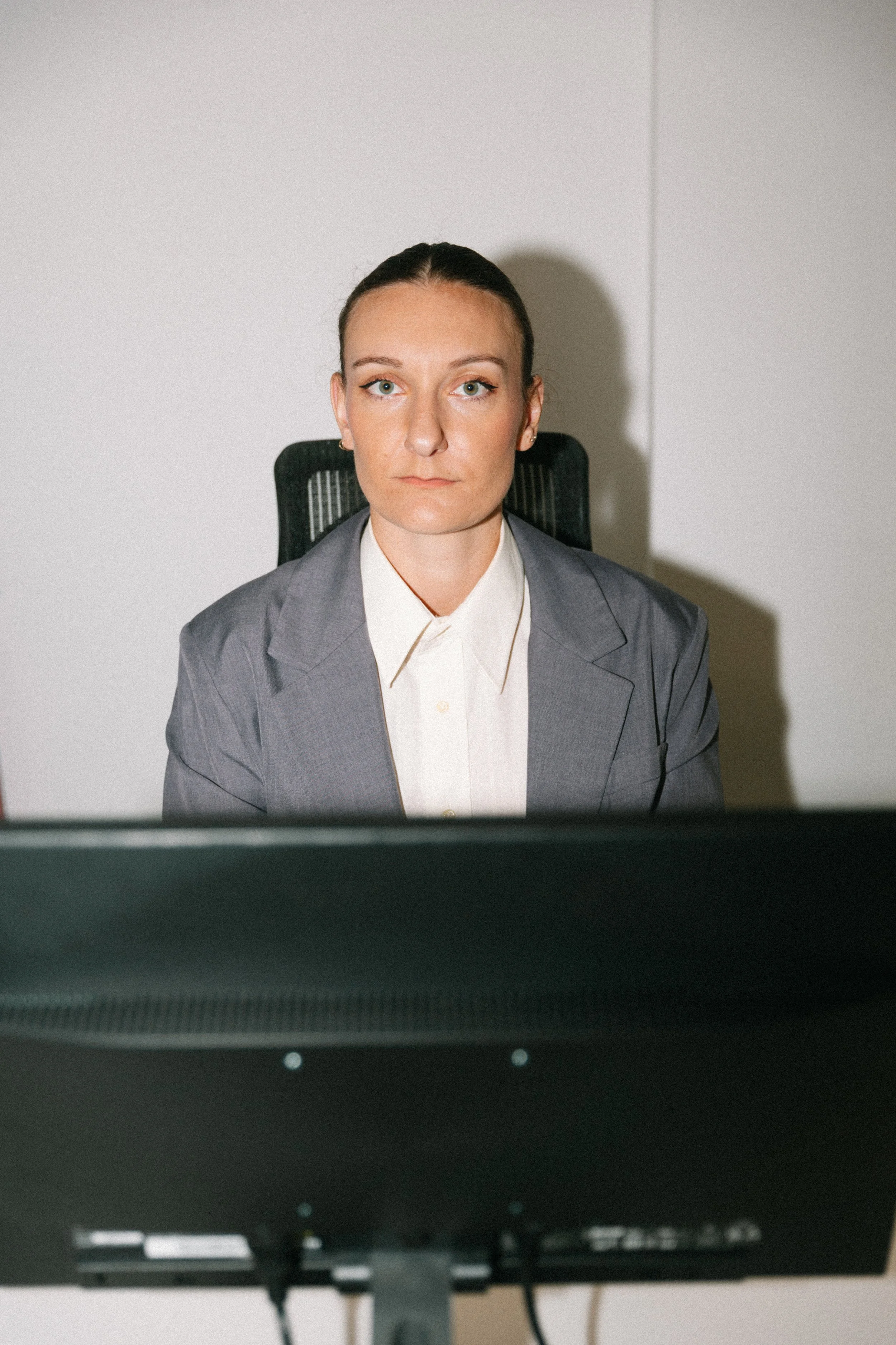 Une femme assise à un bureau, regardant un écran d'ordinateur, en tenue professionnelle gris clair, dans un environnement de bureau moderne.