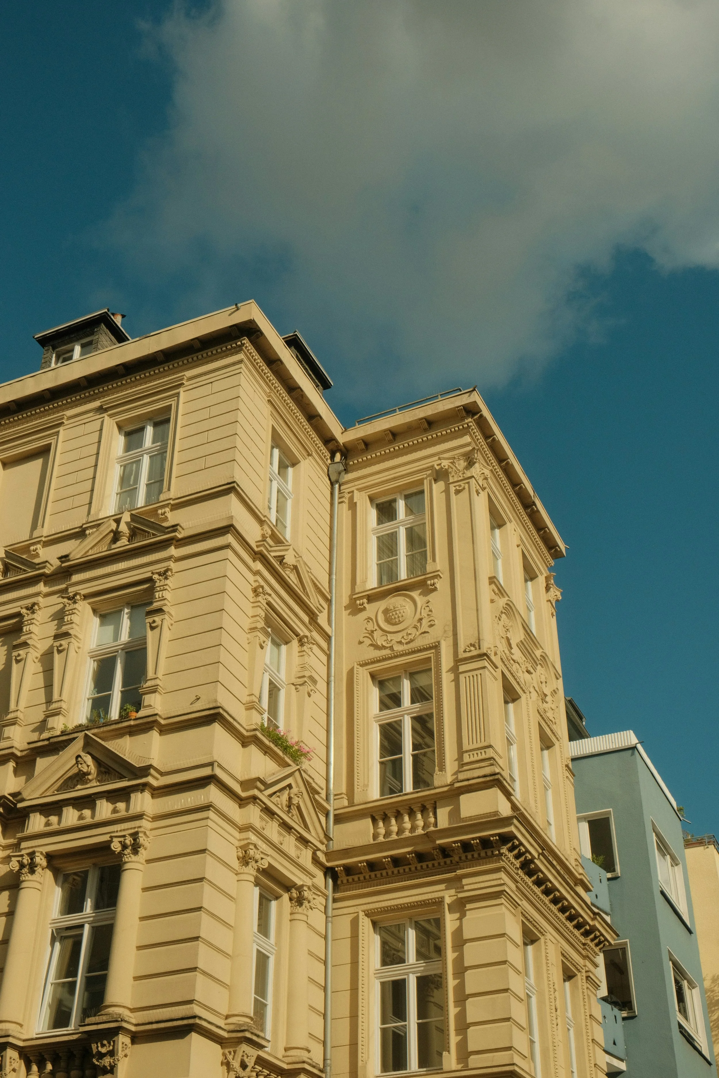 Auf diesem Bild sind die oberen Stockwerke eines historischen, mehrstöckigen, hellgelben Gebäudes mit zahlreichen großen Fenstern und kunstvollen Verzierungen zu sehen, vor einem blauen Himmel mit einigen Wolken.