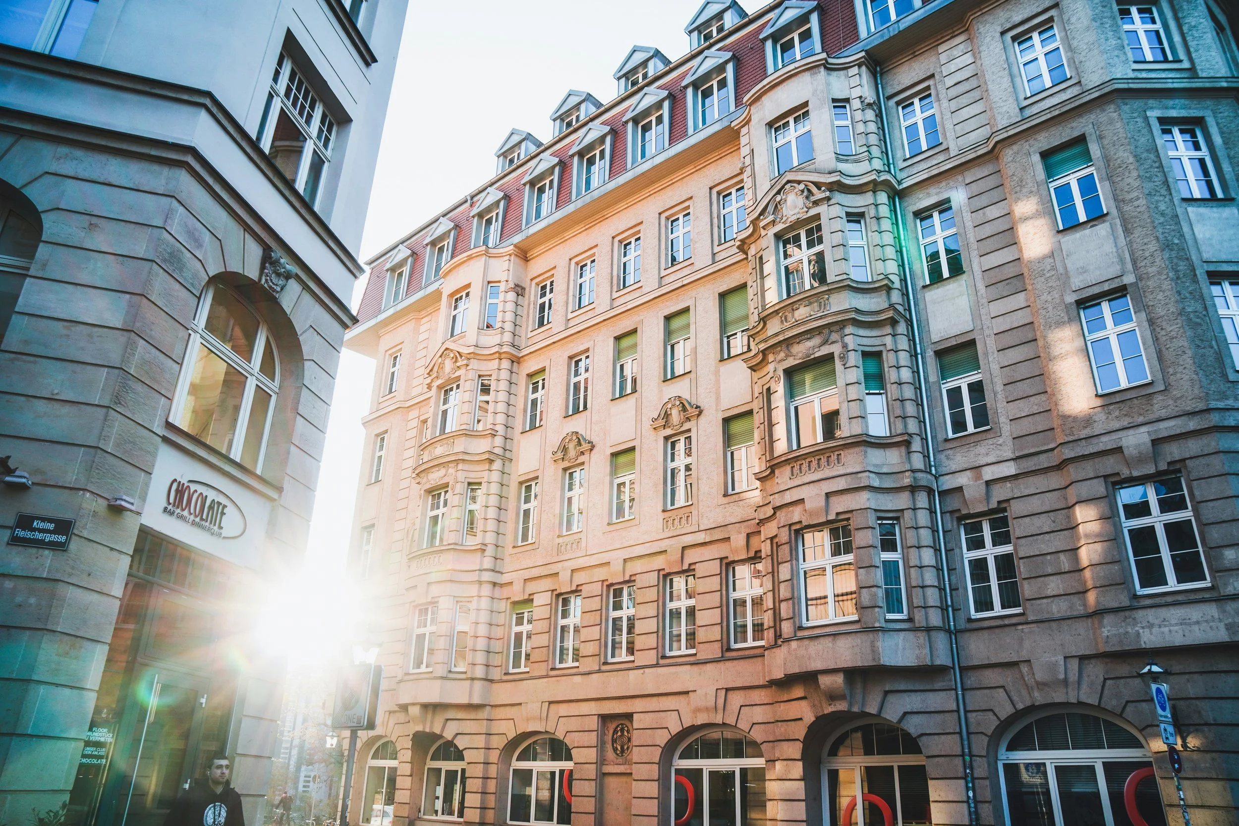Blick auf eine historische Gebäude im Stadtzentrum, im Vordergrund befinden sich Geschäfte und Menschen, es ist Sonnenlicht sichtbar.