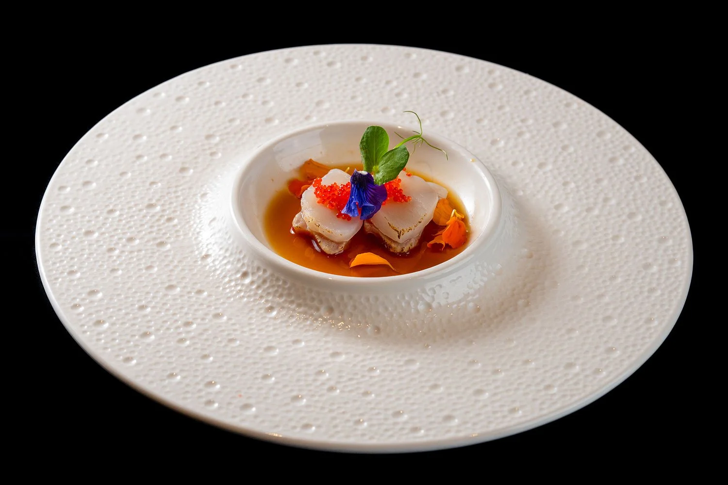 Feines Gericht mit zwei Scheiben Fisch in einer klaren Brühe, dekoriert mit Blümchen, Kräutern und kleinen Torten auf einer weißen, strukturierte Platte.