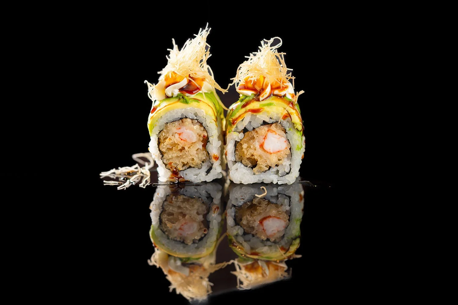 Zwei Sushi-Rollen mit Garnelen, Avocado, Gurke und Topping aus Käsestreifen, Sojasauce, Frühlingszwiebeln und Crispy on Top, auf reflektierendem schwarzen Hintergrund.
