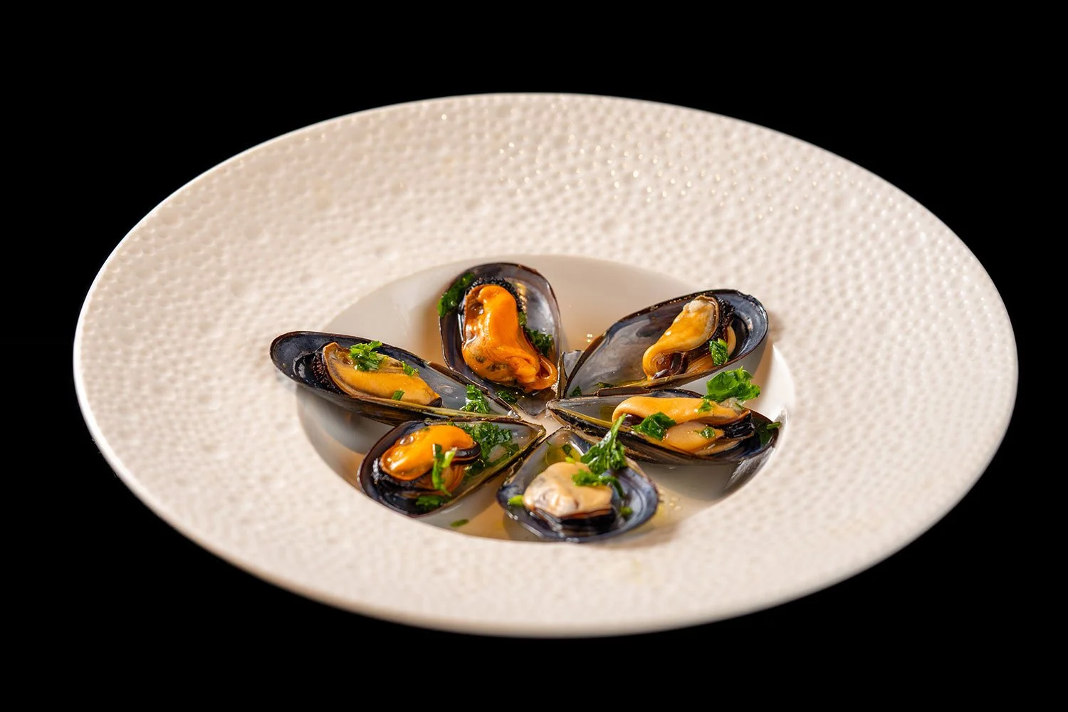 Miesmuscheln auf einem weißen Teller, mit grünen Kräutern garniert, gegen schwarzen Hintergrund.