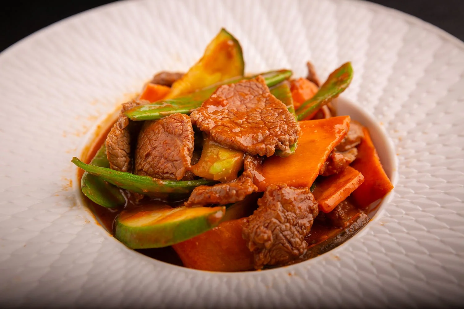 Ein Teller mit asiatischem Rindfleisch-Gemüse-Eintopf, bestehend aus Rindfleischstreifen, Paprika, Zucchini, Karotten und grünen Bohnen in brauner Sauce auf einem weißen, strukturierte Teller.
