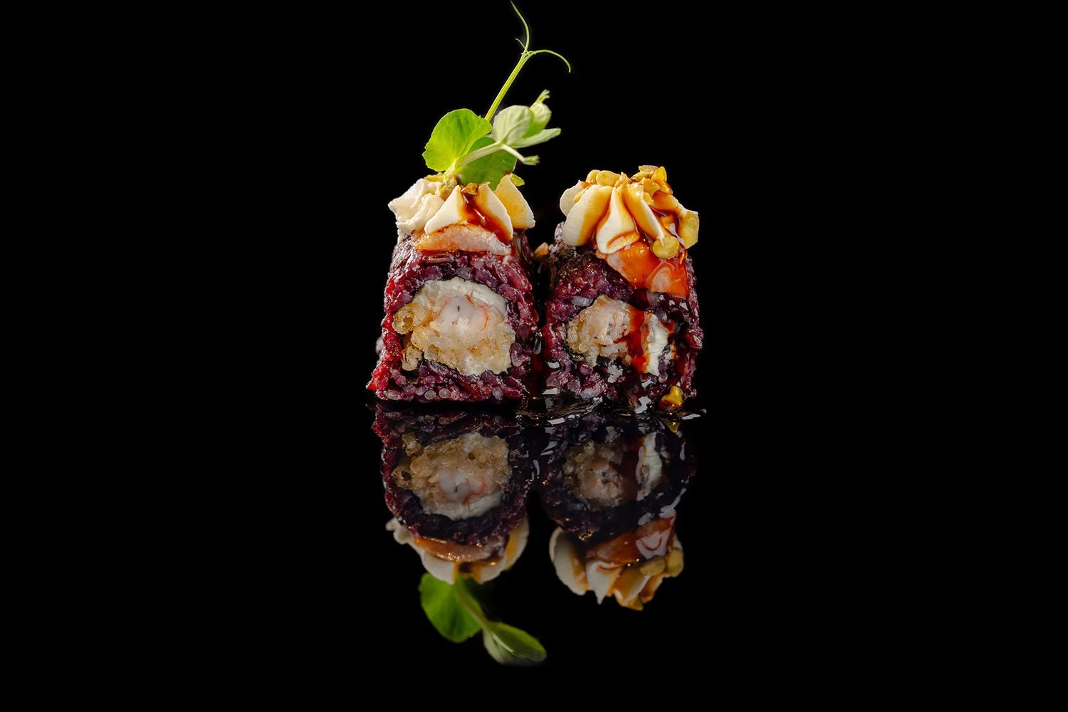 Zwei Sushi-Rollen auf schwarzem Hintergrund, dekoriert mit Sojasauce, Mayo, Nüssen und einem kleinen Blatt, mit Spiegelbild darunter.