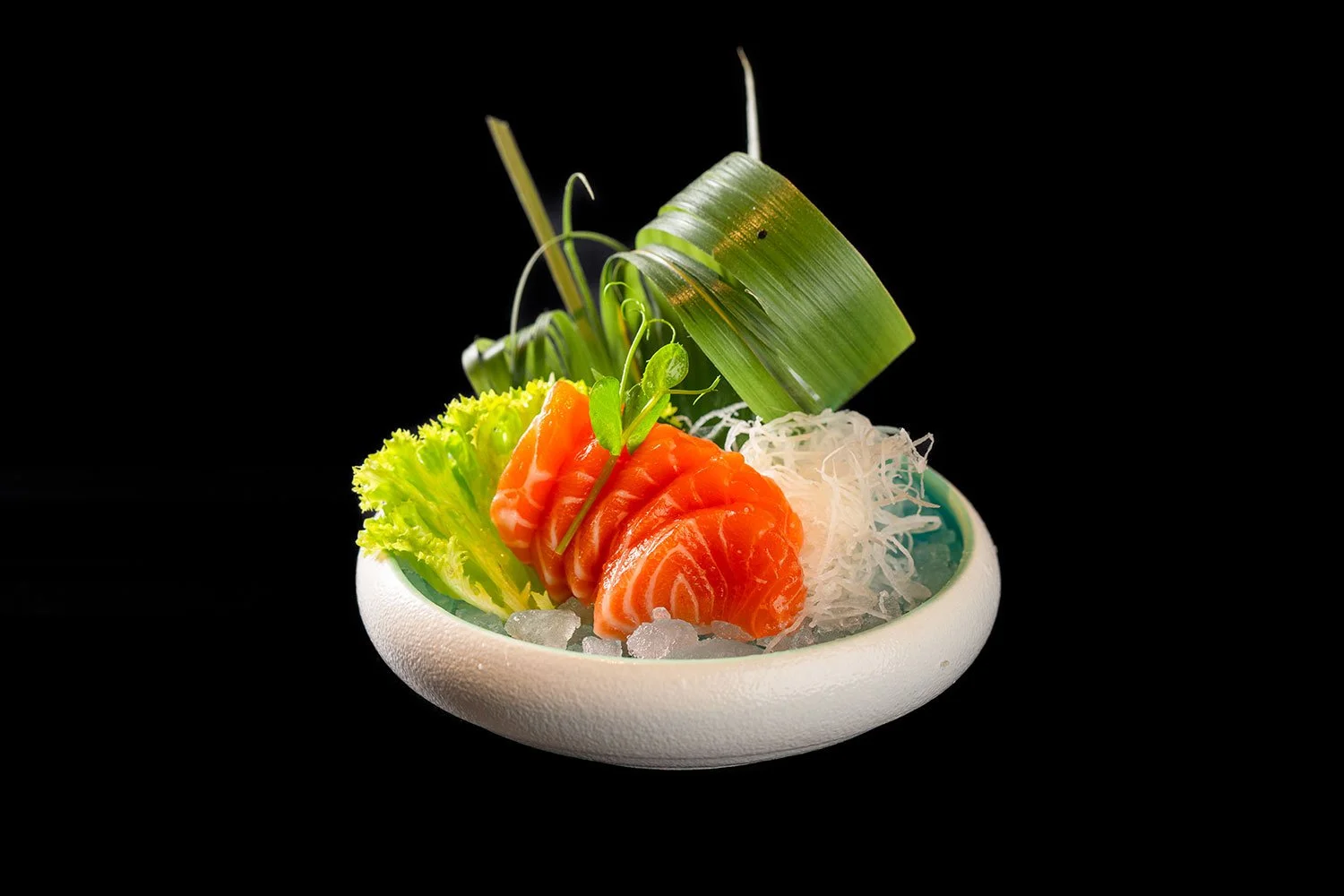 Skandinavischer Lachs Sashimi mit grüner Salatblätter und kunstvoller Dekoration in einer weißen Schale auf schwarzem Hintergrund.