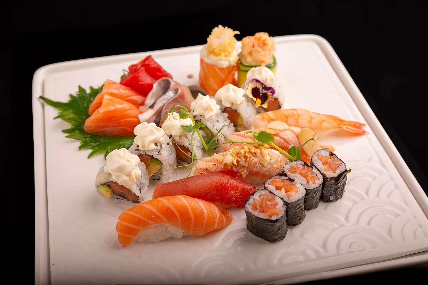 Bunte Sushi-Auswahl auf einem weißen Teller, inklusive Nigiri, Maki, Sashimi und dekorativen Pflanzen, mit Lachs, Garnelen, Thunfisch und Gemüse.