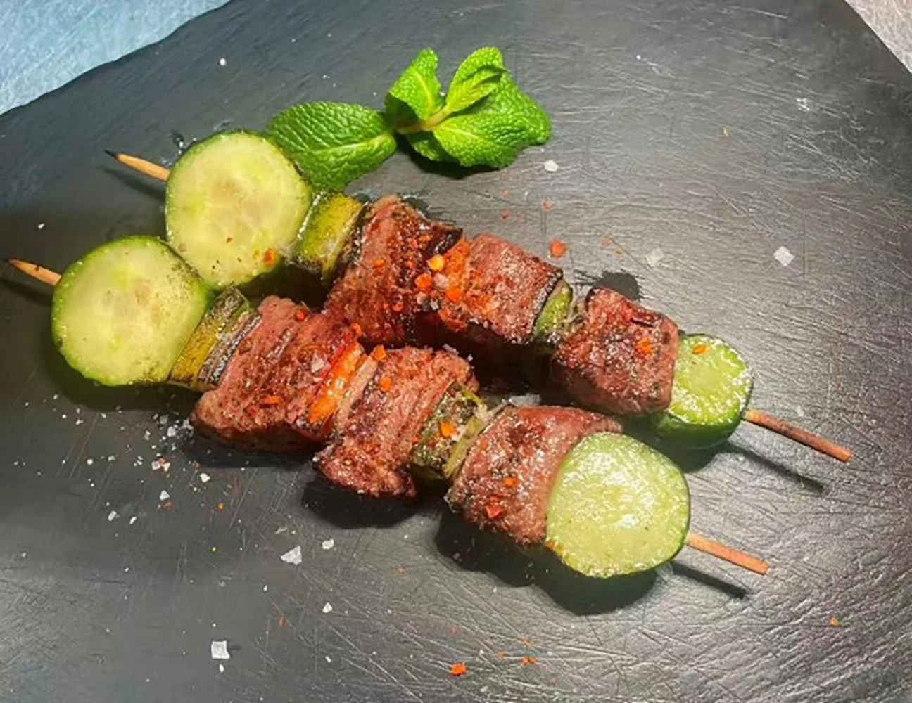 Zwei Spieße mit Rindfleisch, Gurkenscheiben, Gewürzen und frischer Minze auf einem schwarzen Teller.