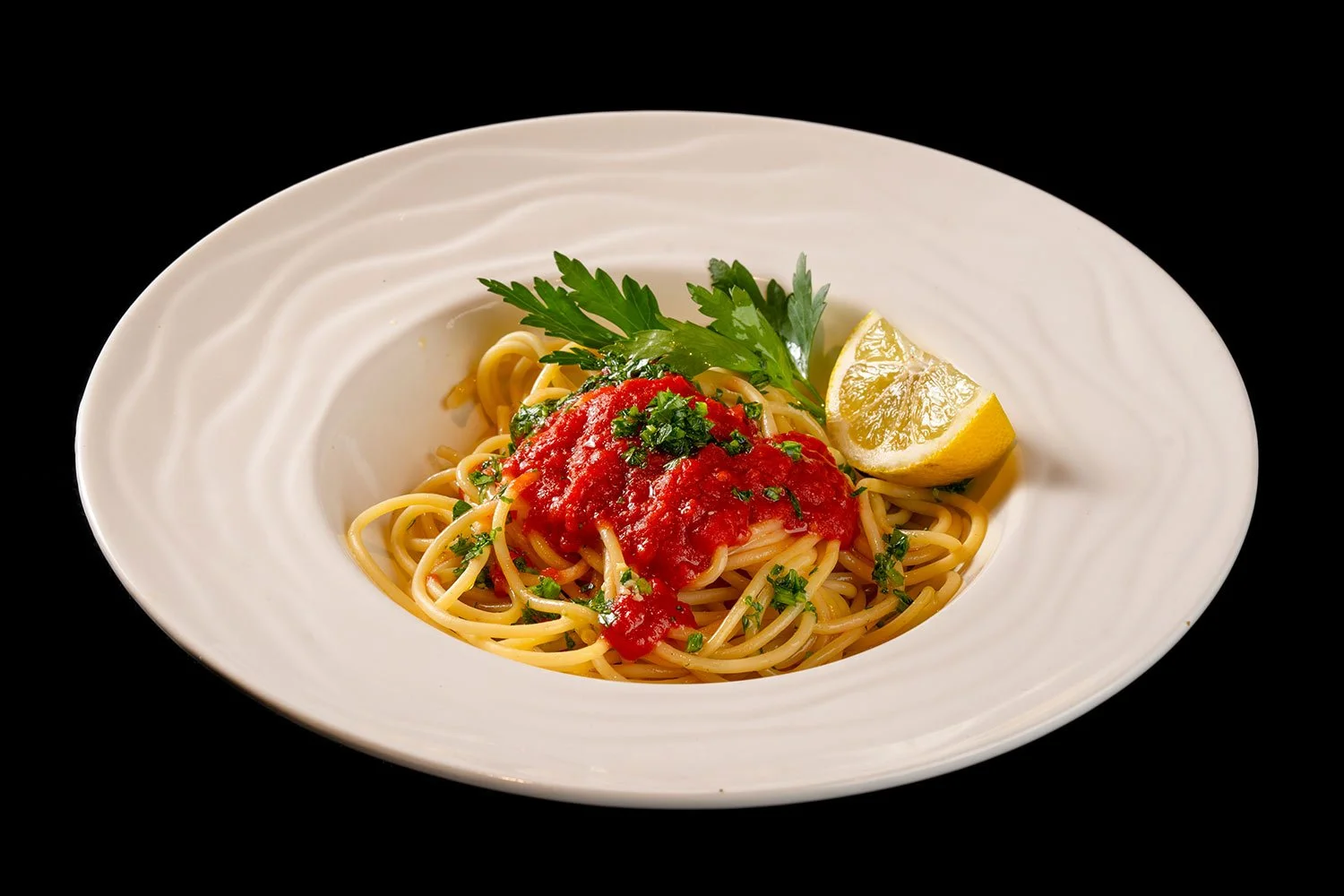 Ein Teller mit Spaghetti, Tomatensauce, frischer Petersilie, einem Zitronenviertel und Kräutern auf einem schwarzen Hintergrund.