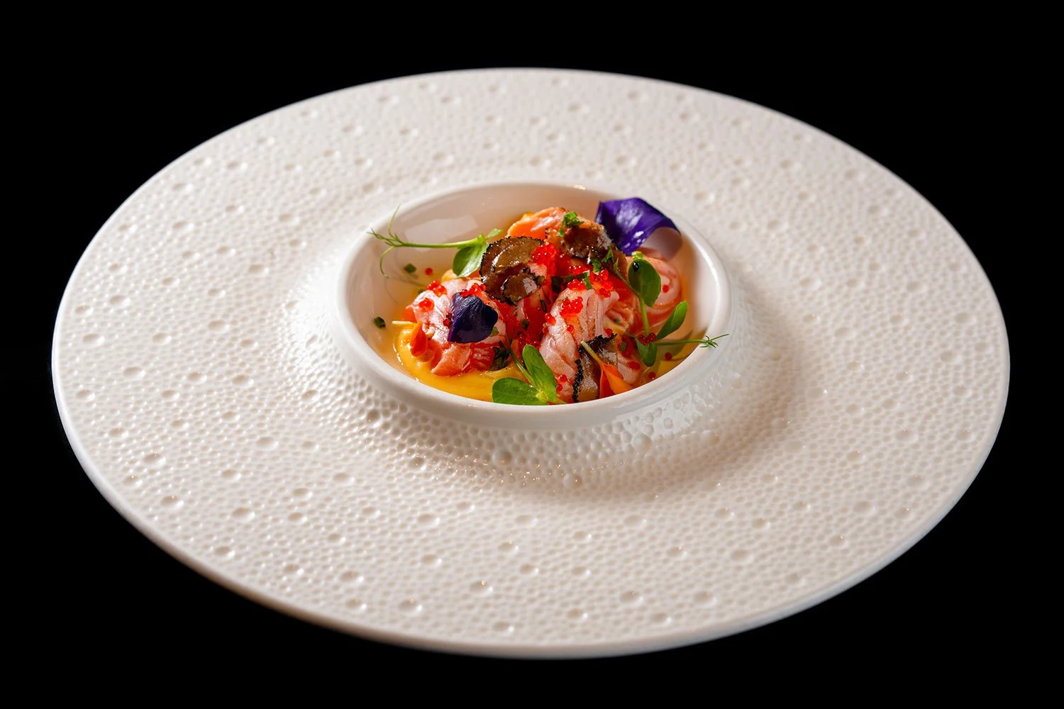 Feines Gericht in einer kleinen weißen Schüssel mit Gemüse, Fisch und Kräutern, serviert auf einem großen weißen Teller mit strukturierter Oberfläche, schwarzer Hintergrund.