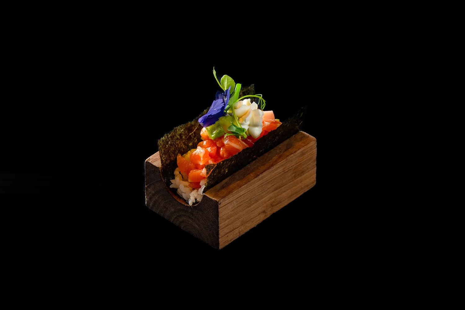 Sushi in einer Holzbox mit Topping aus Tatar, Gurkenscheiben, einer violetten Blüte und essbaren Blumen, auf schwarzem Hintergrund