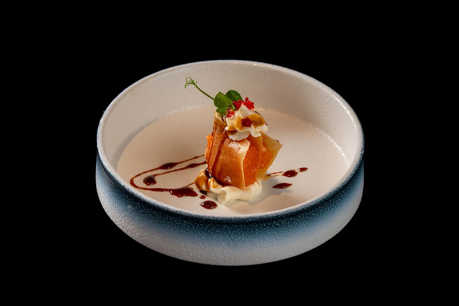 Ein dekoratives Dessert in einer weißen Schüssel mit schwarzem Hintergrund, bestehend aus einer mit Creme gefüllten Garnitur, einem Keks, einer kleinen Menge roter Beeren und einem Minzblatt, garniert mit Schokoladensauce.